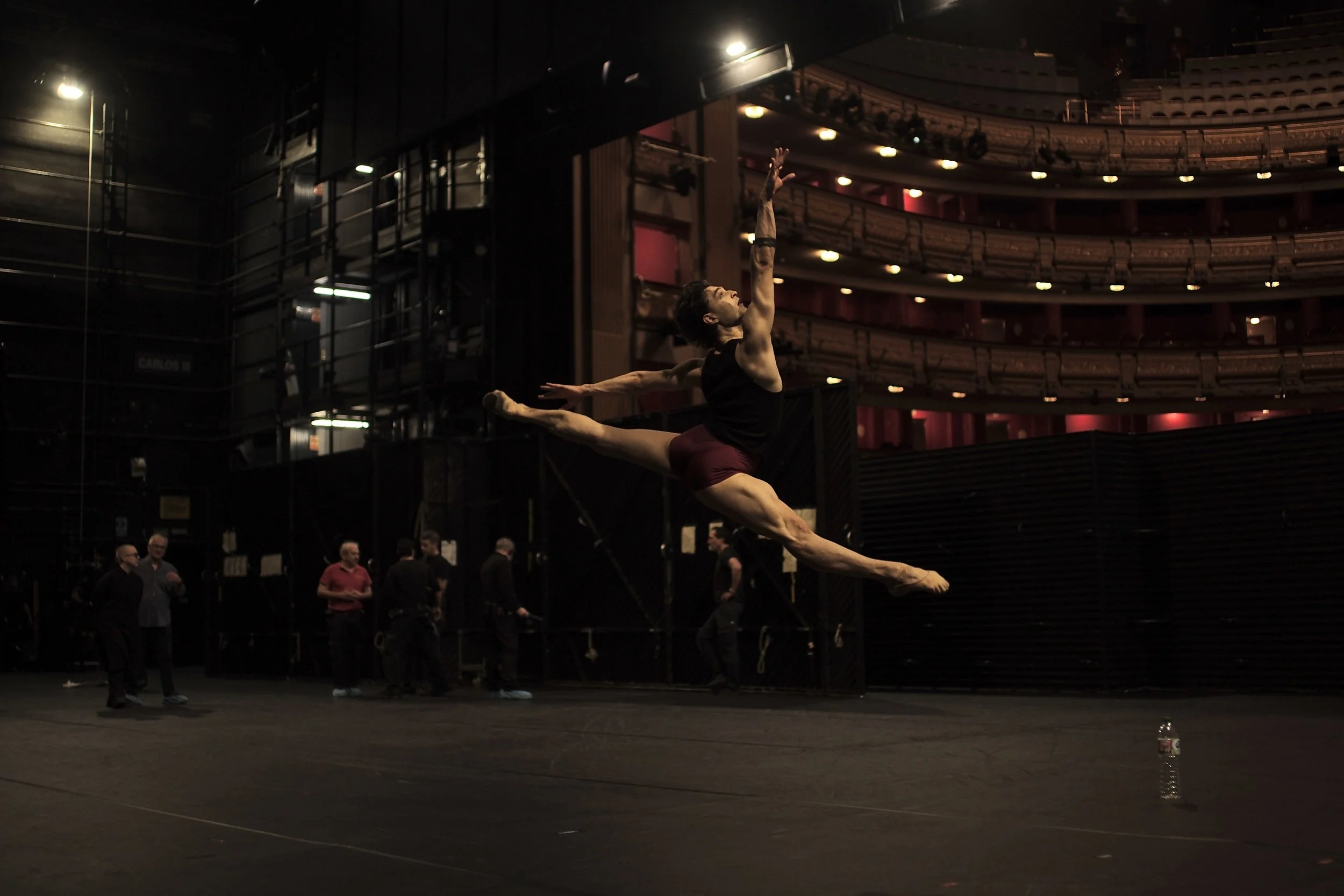 A ballet dancer practicing jumps on stage in a theater with multiple spectators and onlookers around.