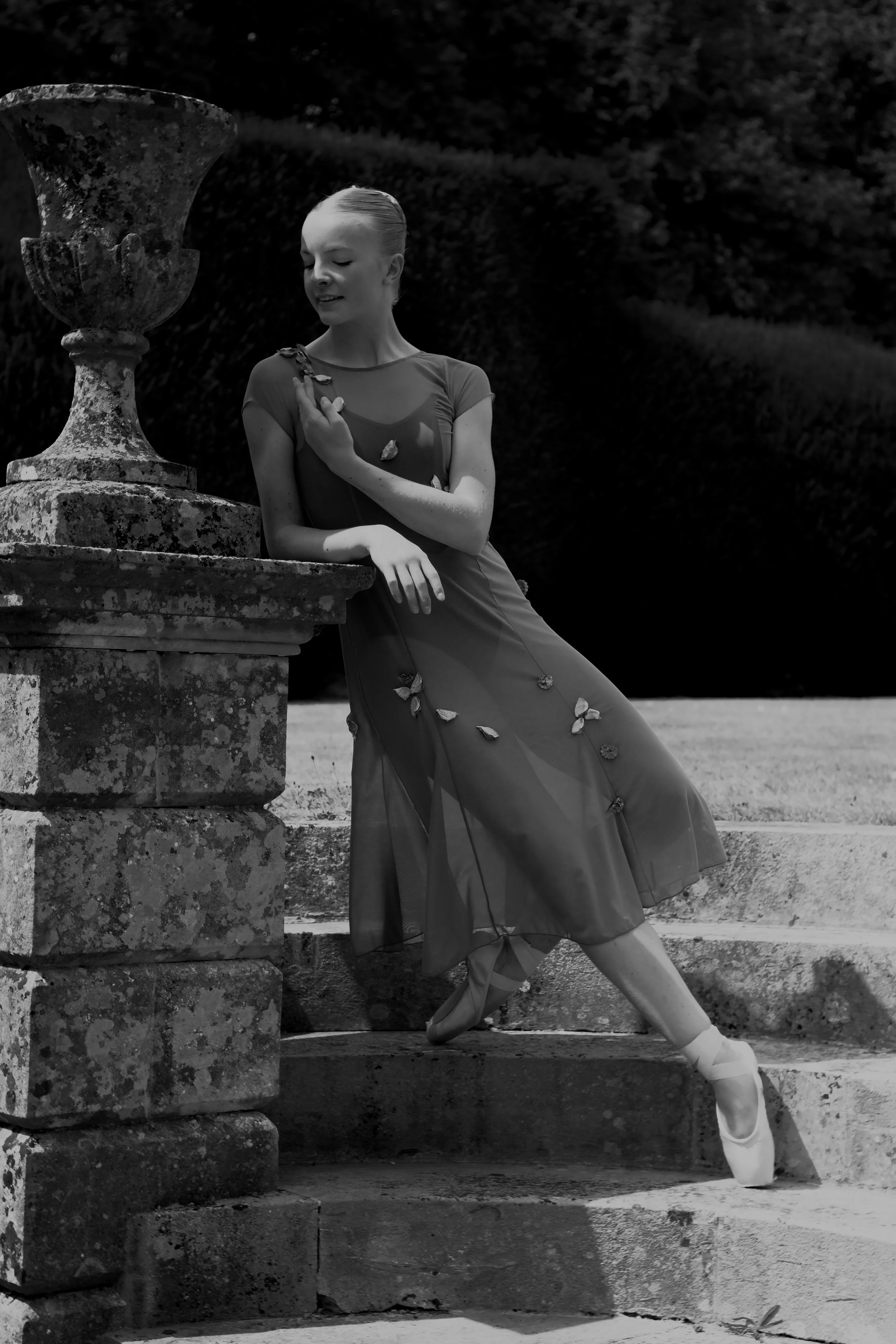 Black-and-white photo of a ballet dancer in a flowing dress, standing on pointe with her leg extended behind her, outdoors near a weathered stone fountain. She gently touches her chest with one hand and gazes down with a serene expression.