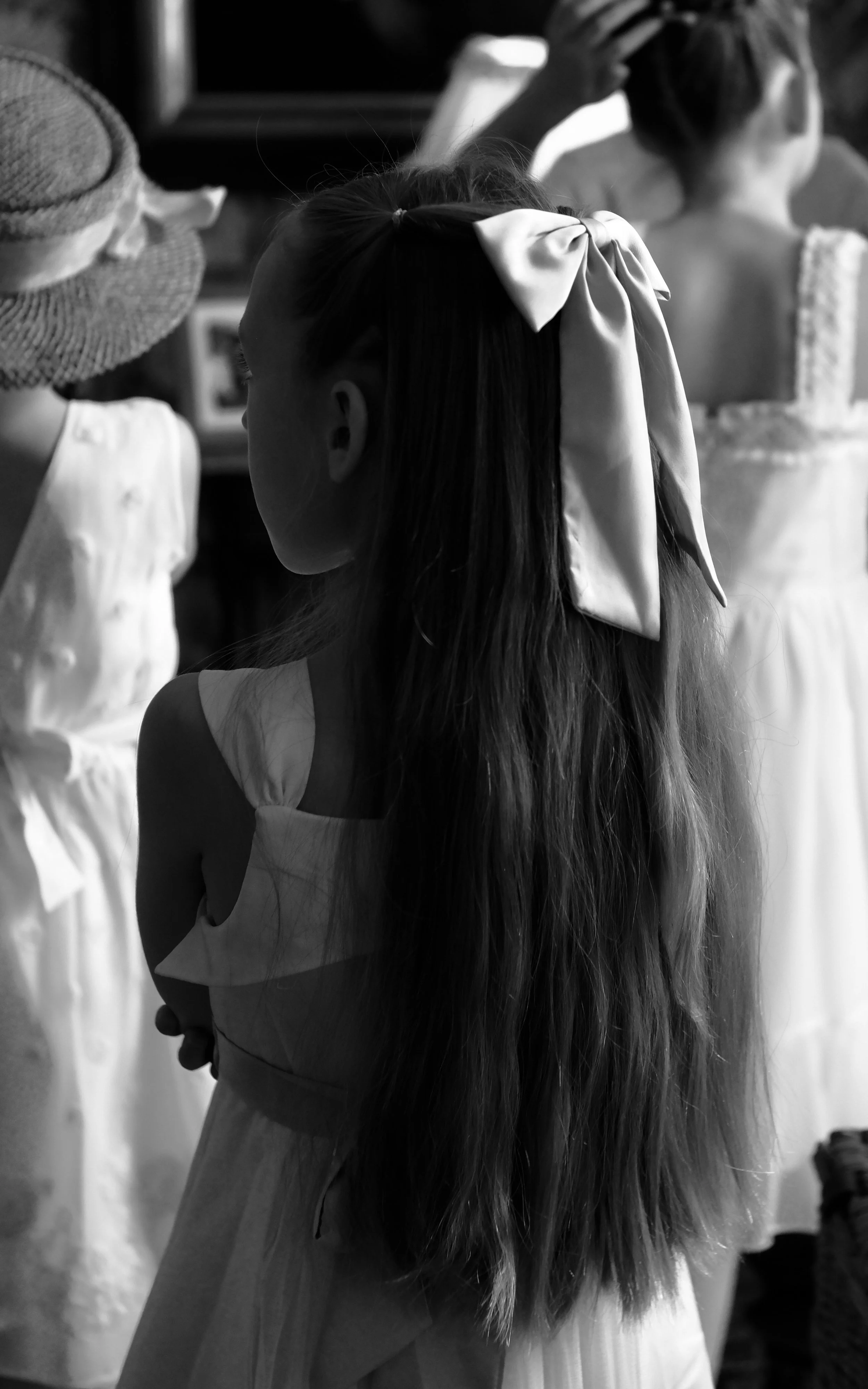 A young girl with long hair tied with a large white bow, wearing a sleeveless dress, standing in front of a display with mannequins dressed in fancy outfits.