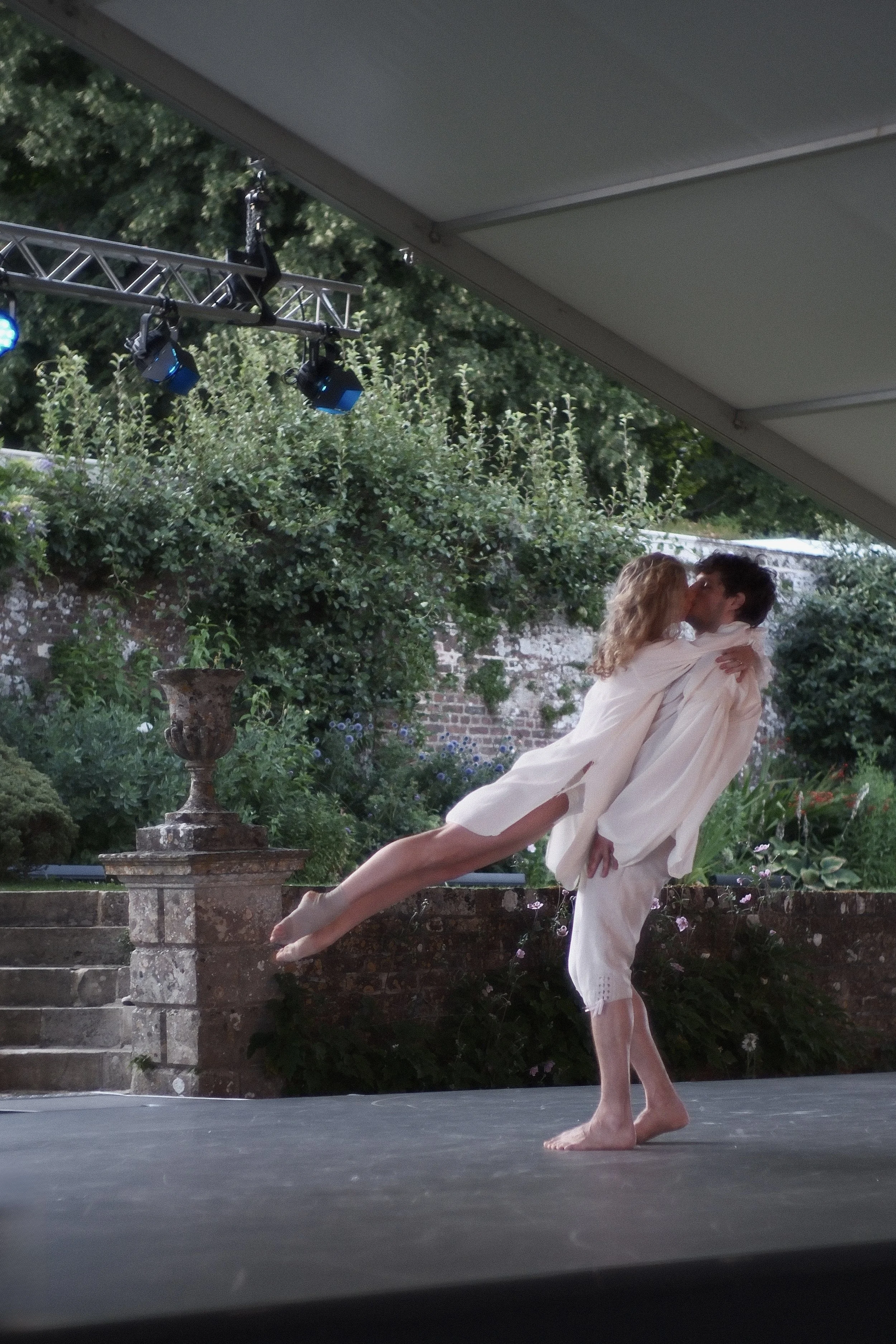 Two ballet dancers, a man and a woman, performing a lift outdoors on a stage under a tent, with greenery and brick steps in the background.