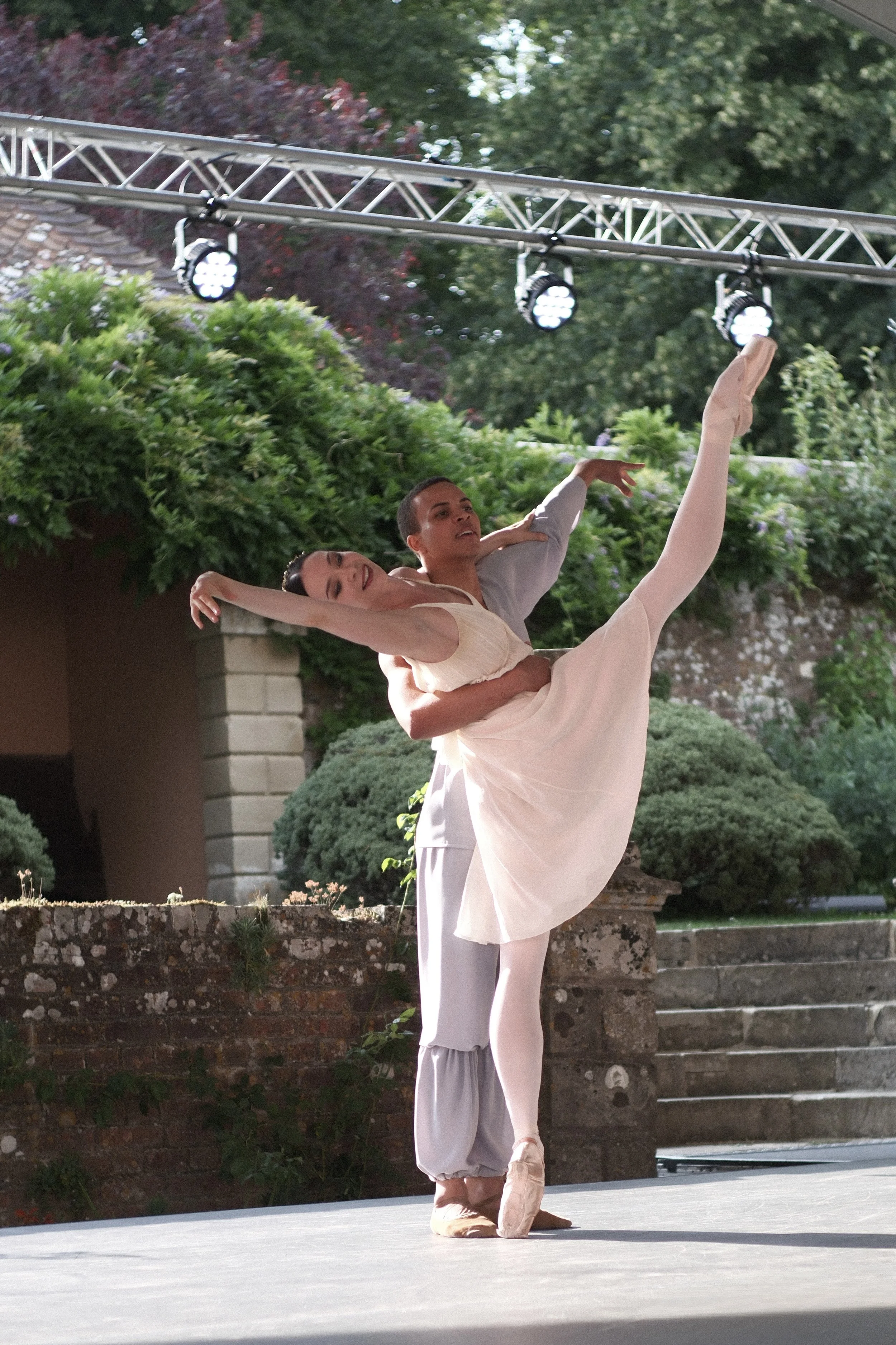 Two ballet dancers performing outdoors, with the male dancer lifting the female dancer in an arabesque position. They are dressed in light-colored costumes, and are on a stage with greenery and steps in the background.
