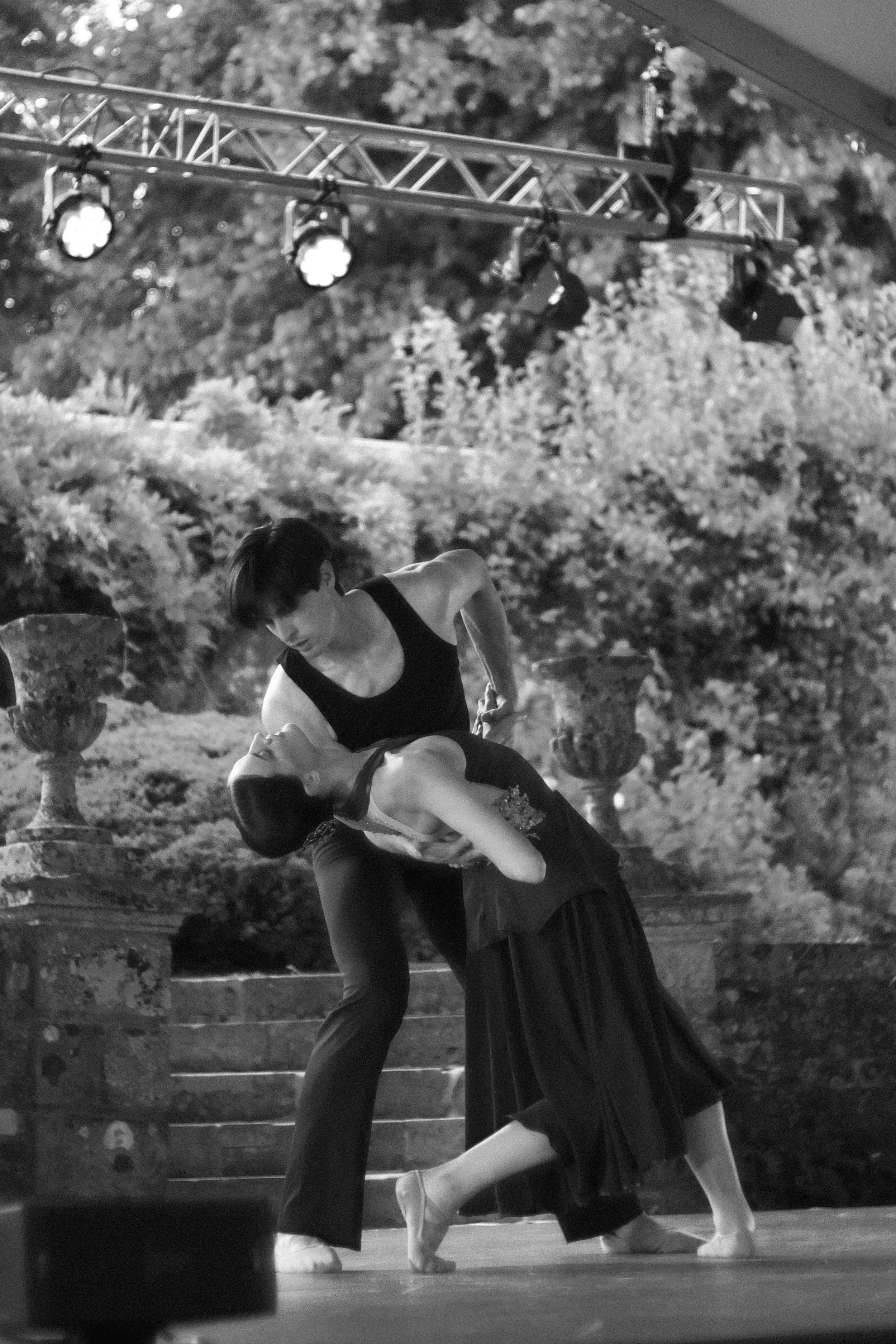 Two ballet dancers performing on stage, one in a black dress and the other in a tutu, holding each other, surrounded by stage lights and trees in the background.