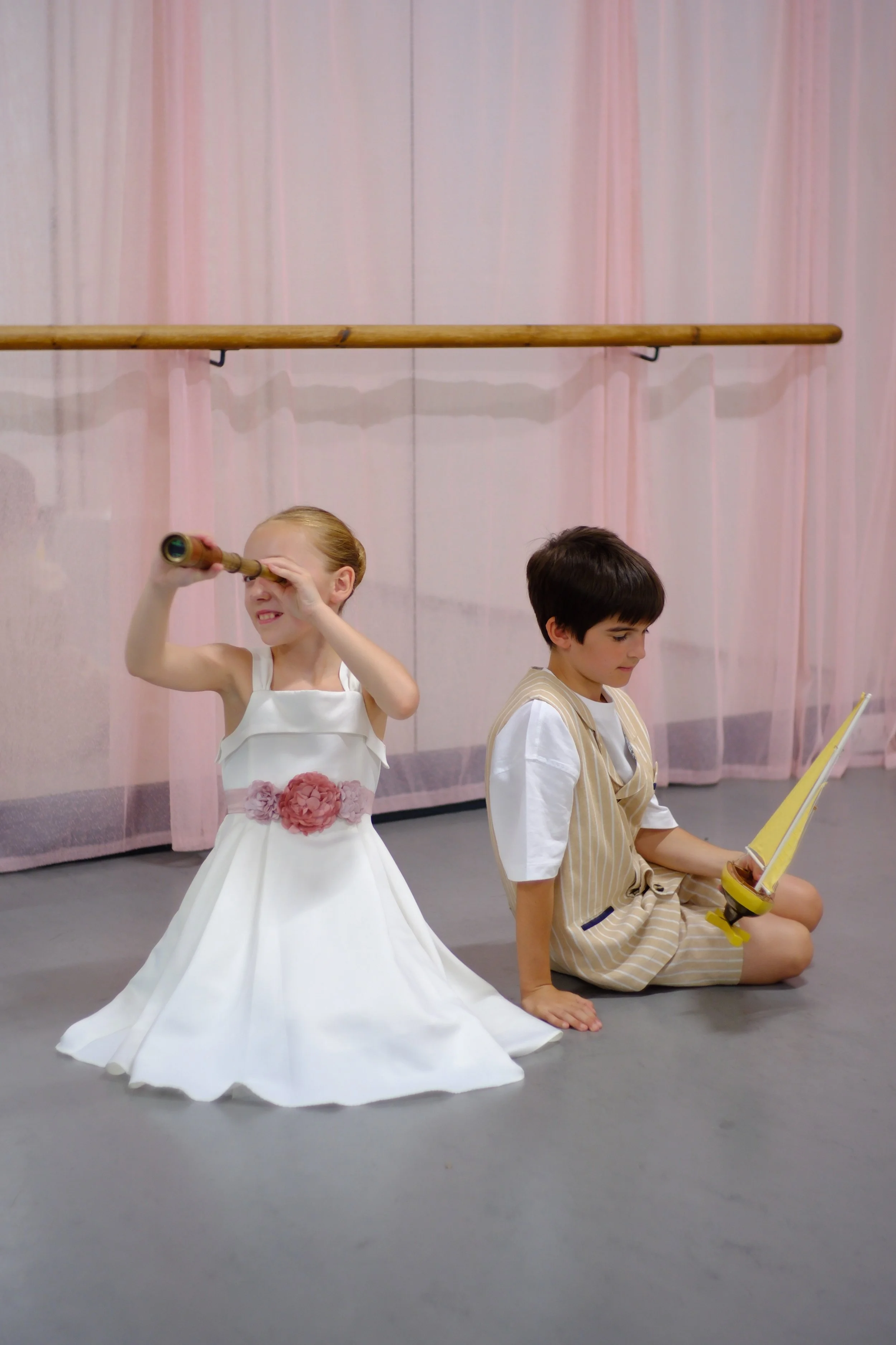 Two children dressed as a princess and a knight sitting on the floor in a dance studio with pink curtains and a ballet barre in the background. The princess is smiling and looking through a telescope, while the knight is reading a book.