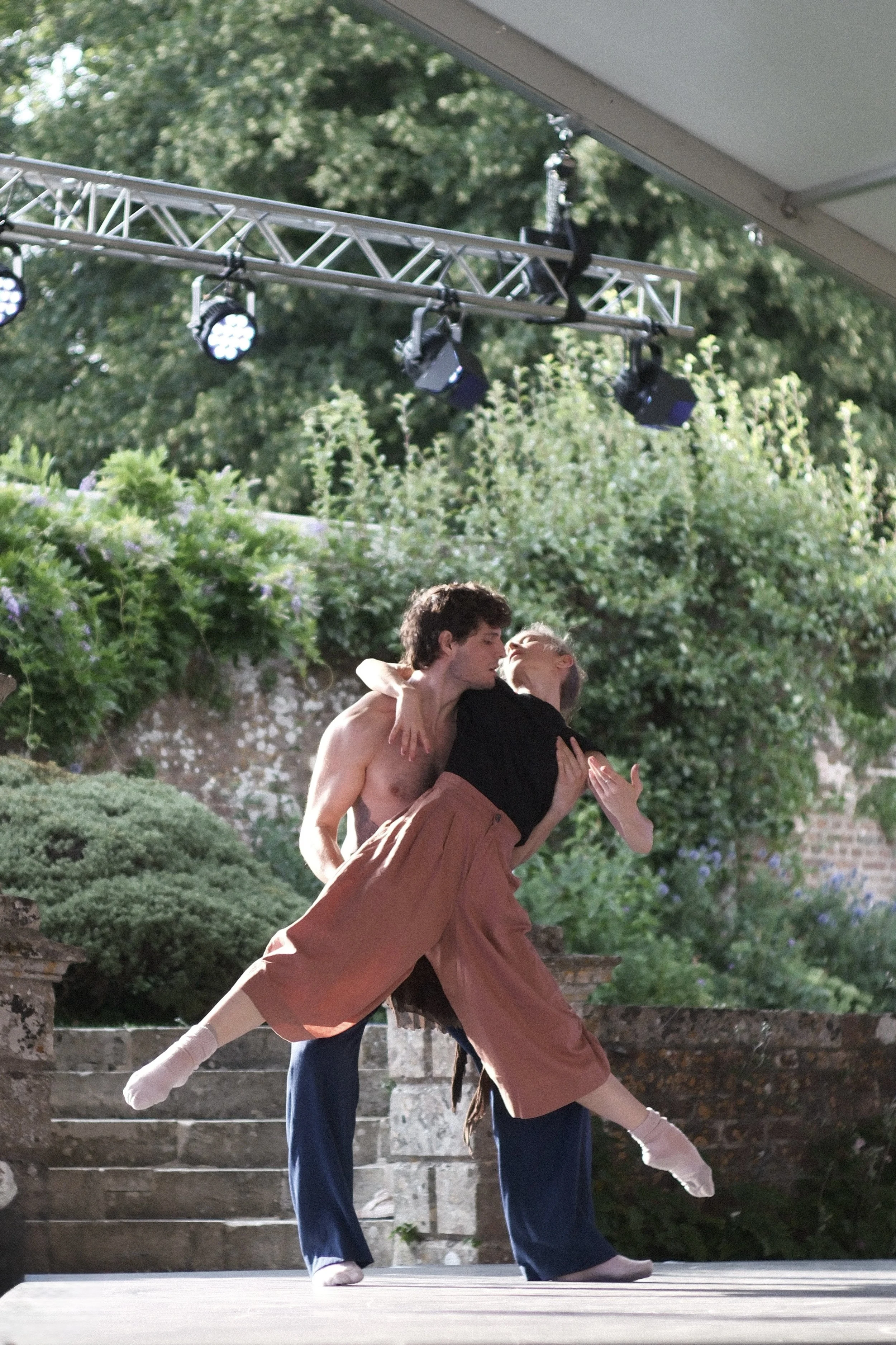 Two ballet dancers performing outdoors, one carrying the other in a dance pose, with a backdrop of greenery and stone stairs.