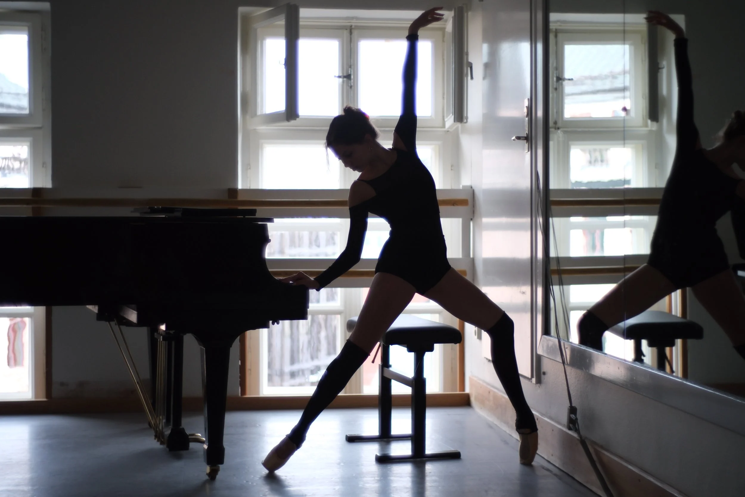 Ballet dancer practicing at a piano in front of large windows with sunlight streaming in.