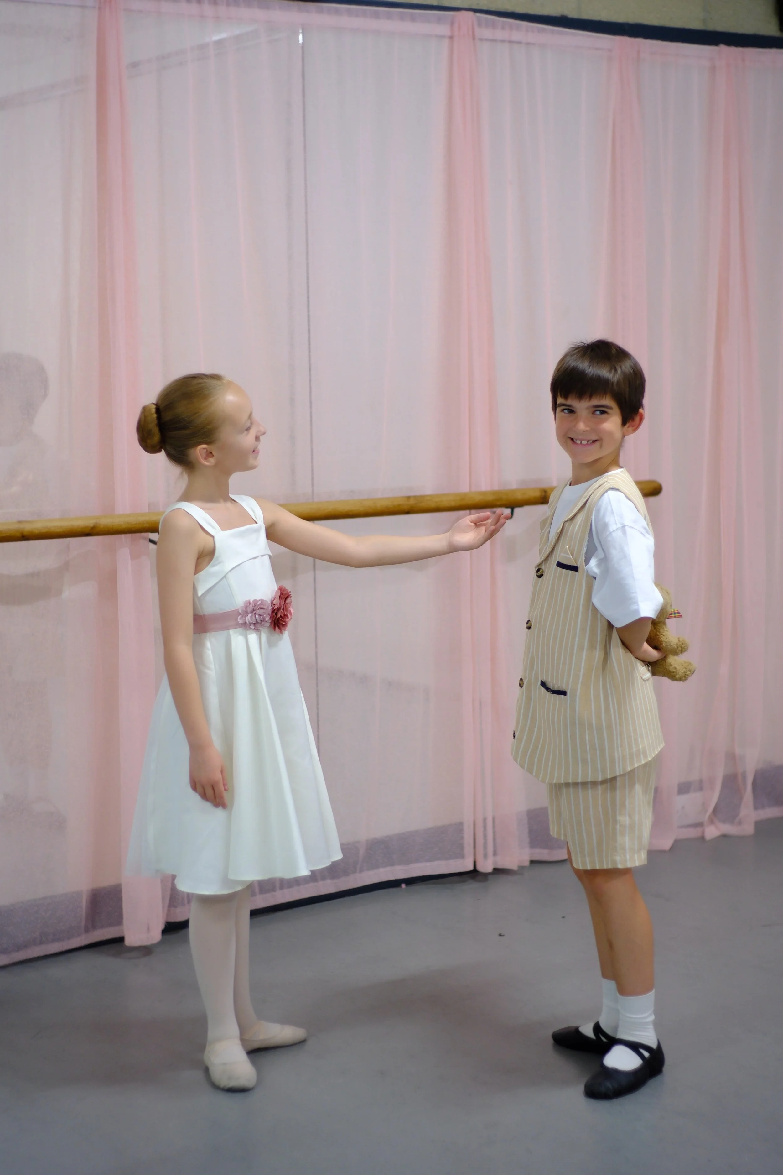 Two young children, a girl and a boy, dressed in ballet outfits, are standing in a dance studio with pink curtains and a ballet barre. The girl has her hair in a bun, and the boy is smiling at the camera, holding a teddy bear behind his back.