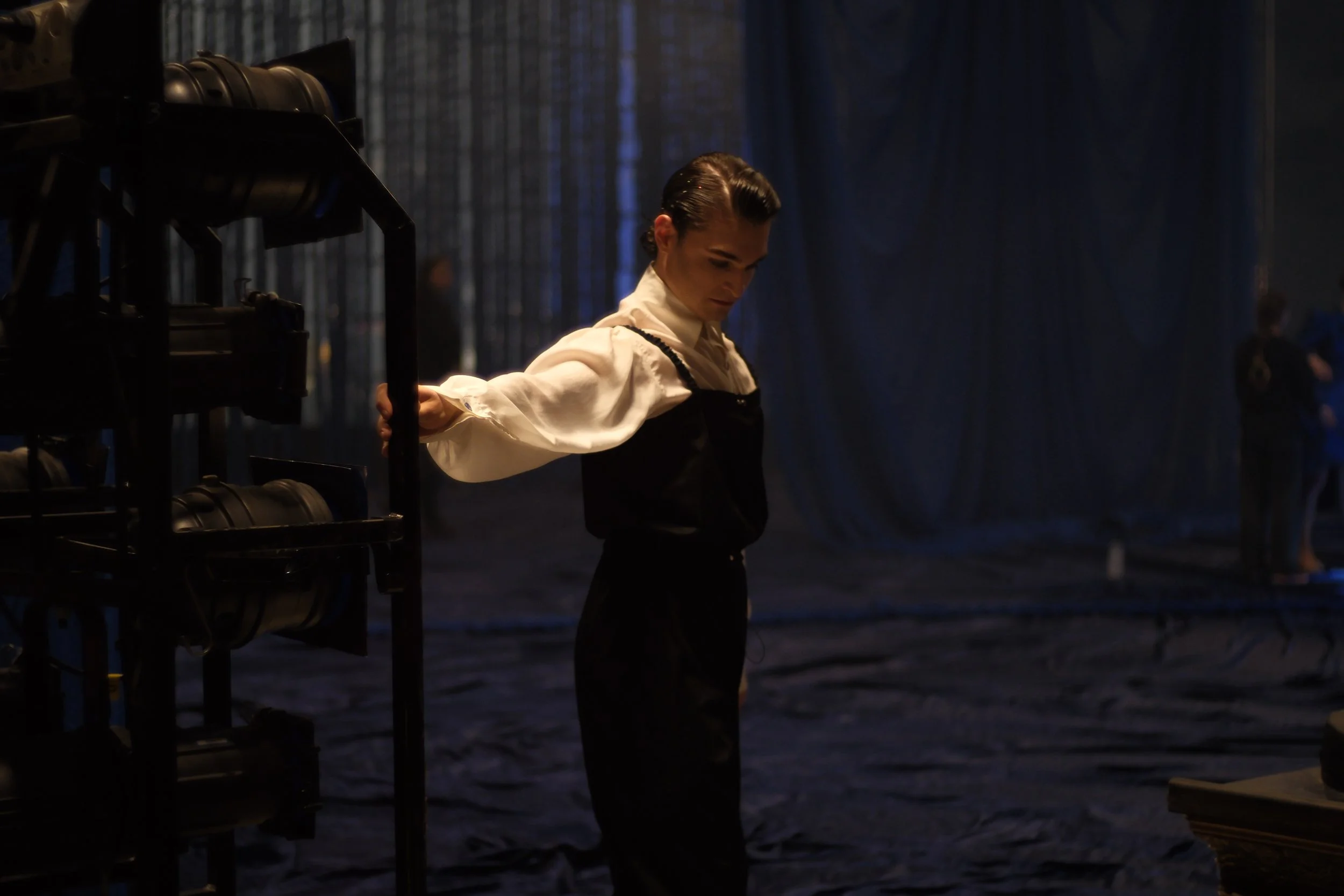 A woman with vintage hairstyle, dressed in white blouse and black apron, stands backstage behind metal shelves filled with stage lights, with blue stage curtains in the background, in a dimly lit theater.