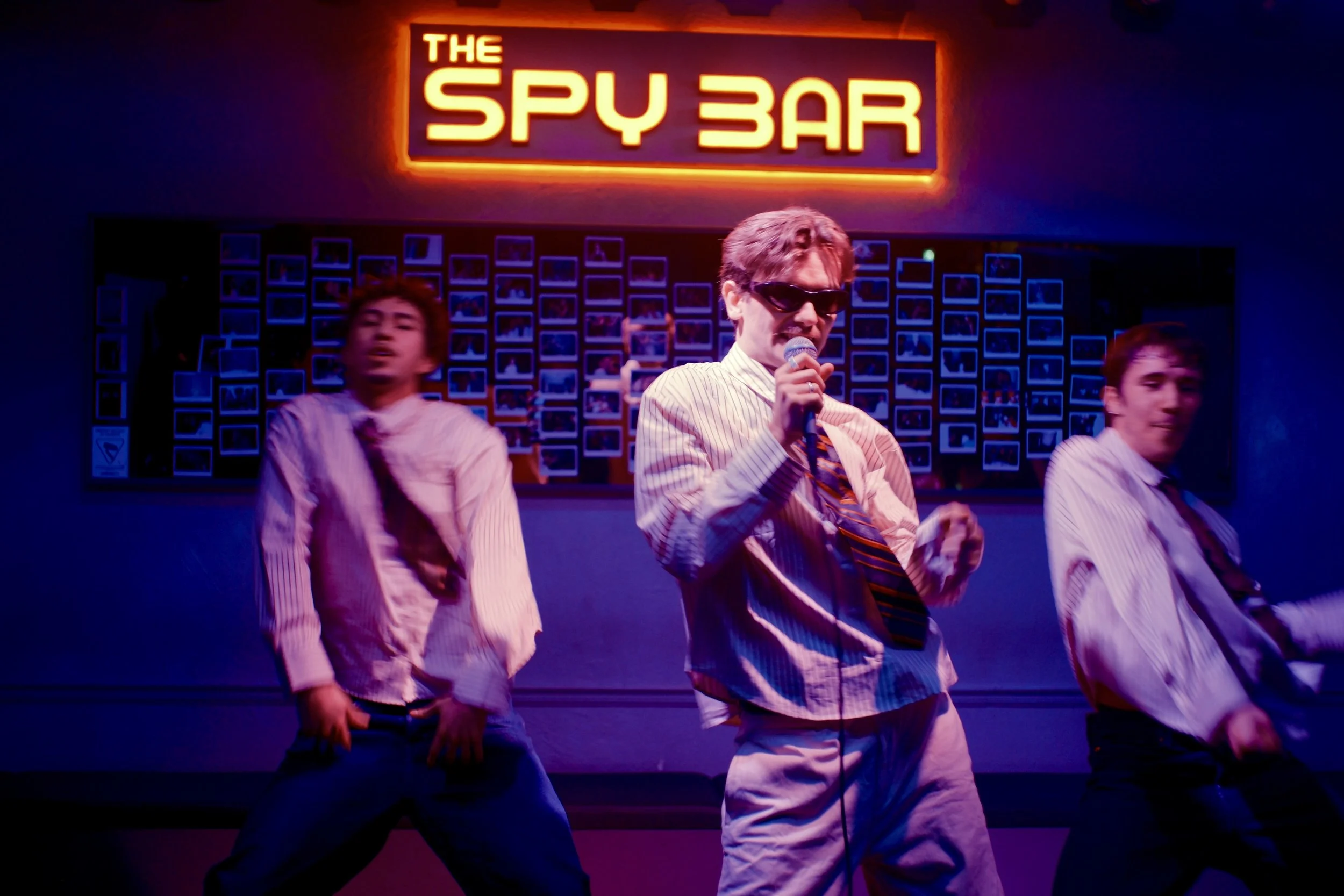 Three men dancing on stage in front of a neon sign that reads 'THE SPY BAR.' The man in the center is wearing sunglasses and a striped shirt, holding a microphone, and the other two are wearing striped shirts as well. The background features a wall w