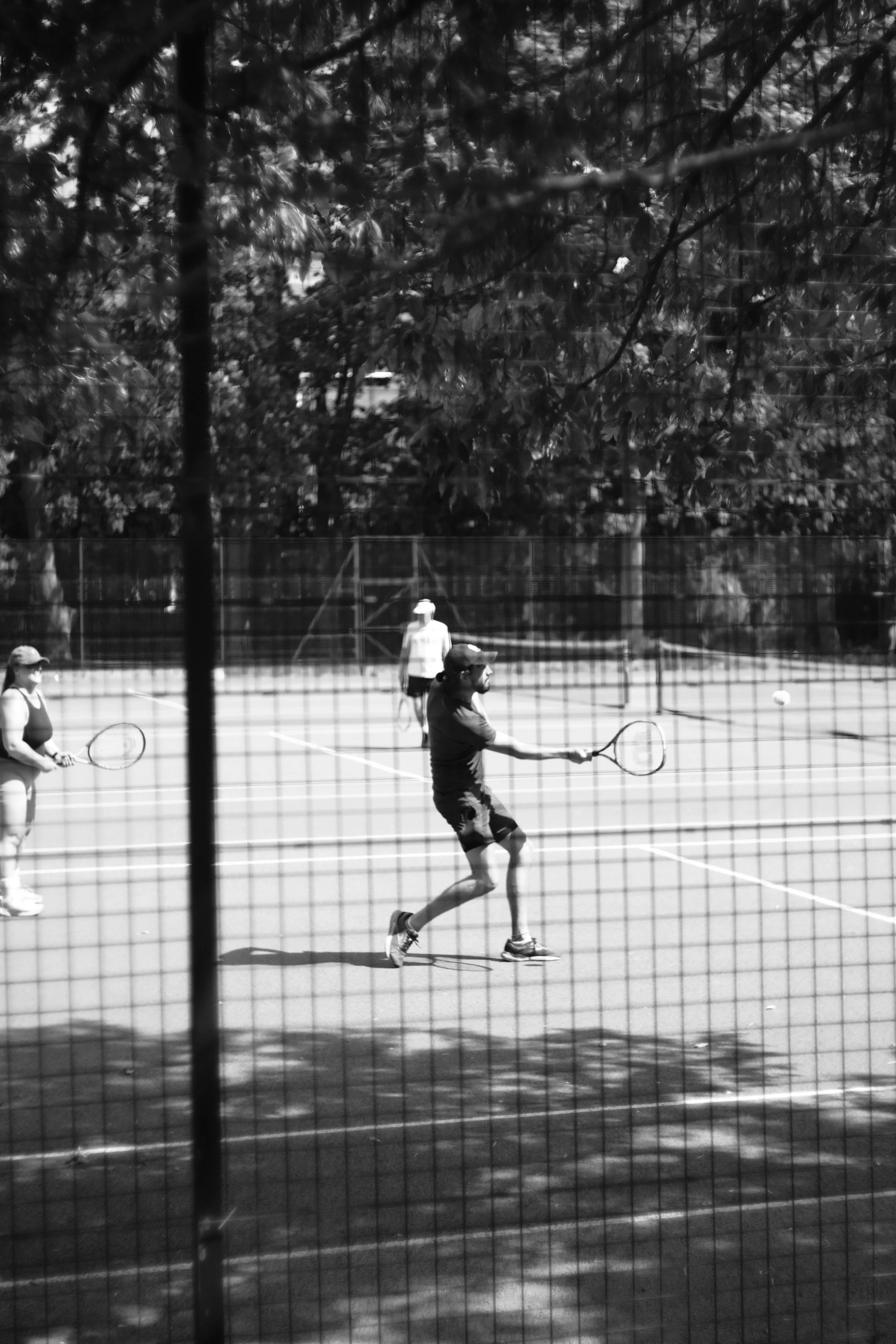 Black and white photo of tennis players on a court, one in focus hitting a ball, others observing. Trees and a fence in background.