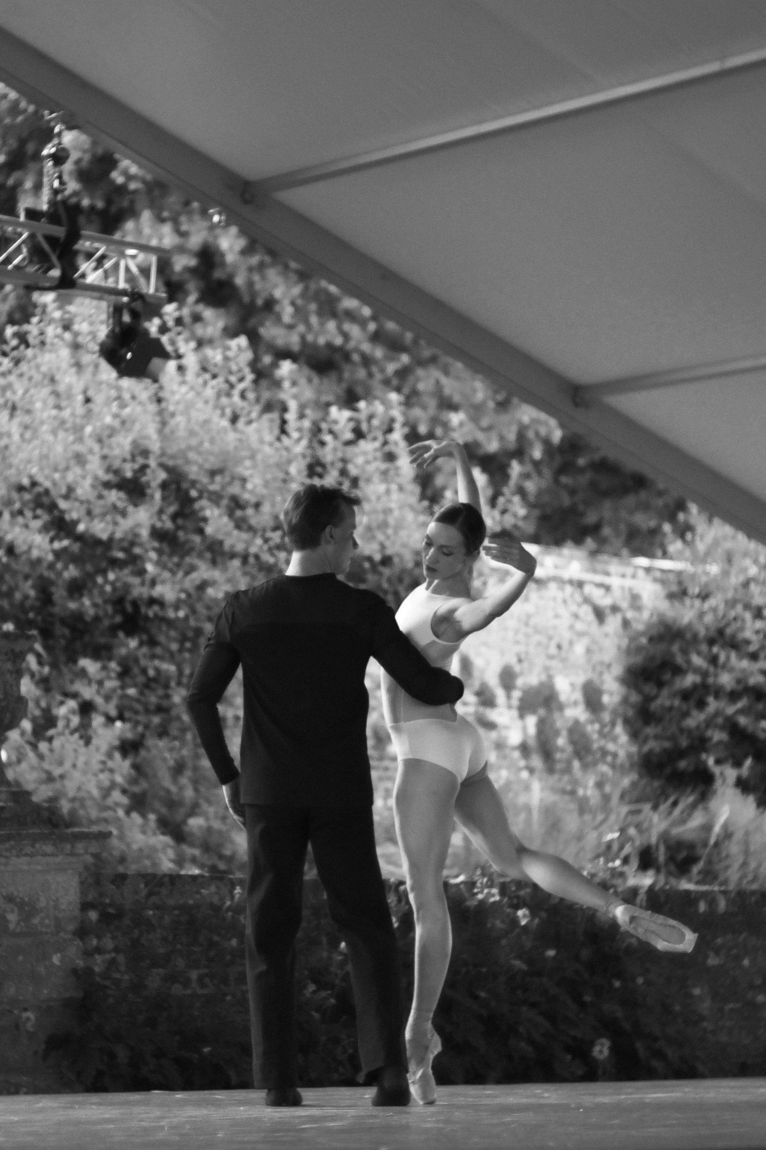 A male ballet dancer and female ballet dancer practicing together outdoors, with trees in the background. The female dancer is in a ballet pose with one arm raised and the other extended behind her, wearing a leotard and tights. The male dancer, in a