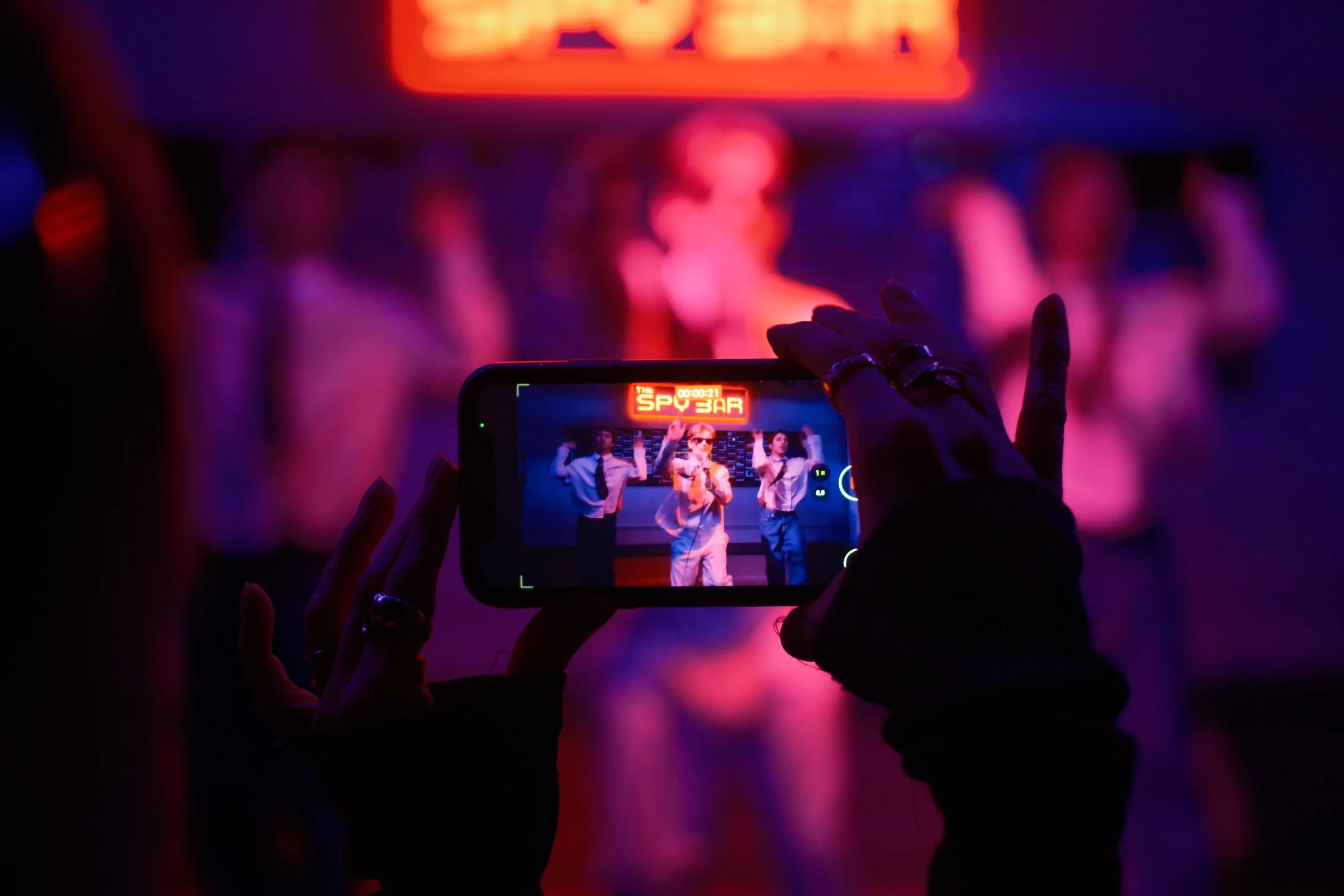 Person taking a photo of a musical performance on stage with a smartphone in a dimly lit venue featuring vibrant purple and red lighting.