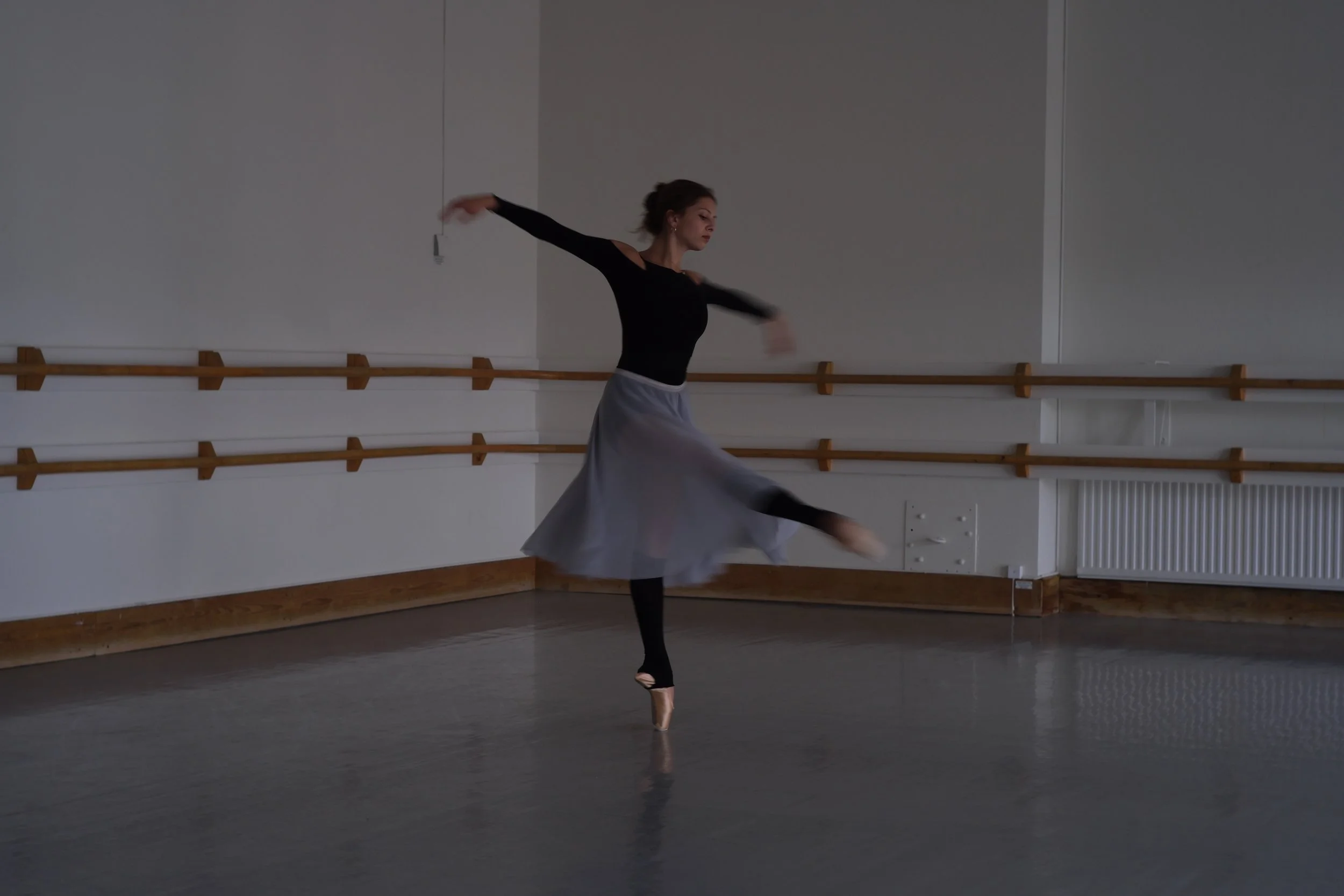 A ballet dancer practicing in a dance studio with wooden ballet barres on the wall. The dancer is performing a move on pointe, with her arms extended and one leg lifted in the air. She is wearing a black top, a gray skirt, and black tights.