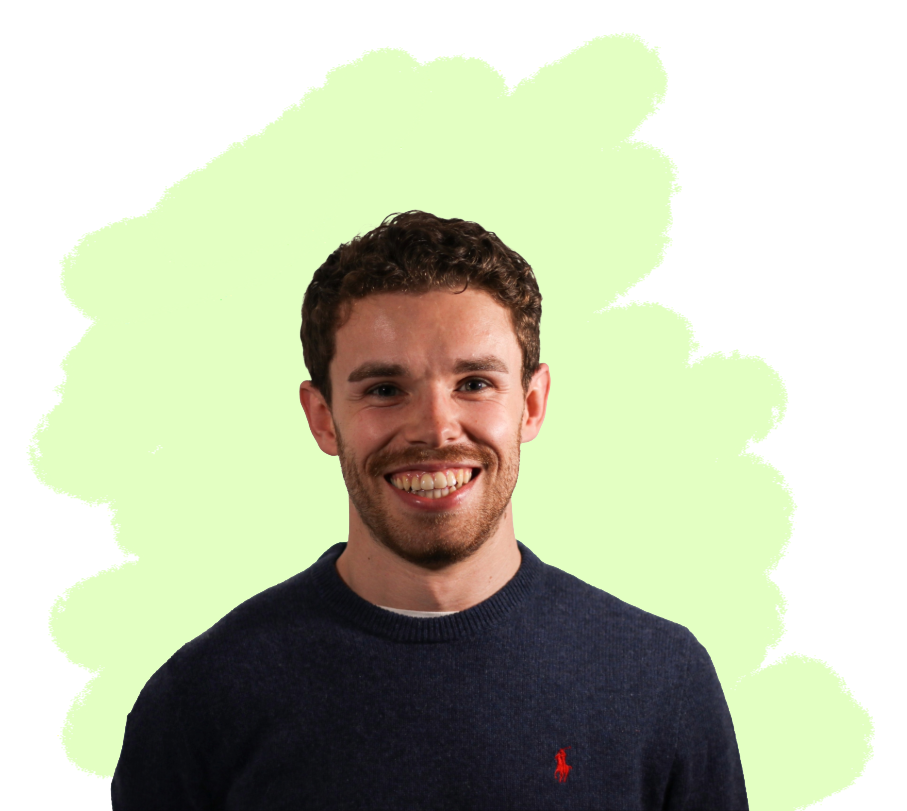 A smiling young man with short curly brown hair and a beard, wearing a dark navy blue sweater with a small red logo on the chest, standing against a light green background.