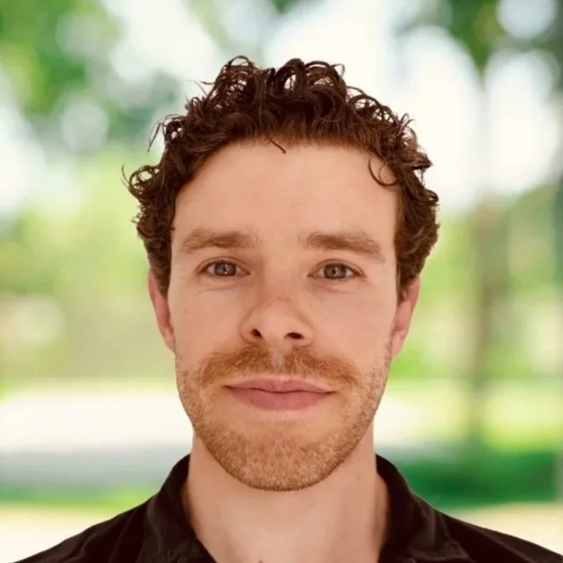 Close-up portrait of a man (Collum) with curly brown hair, light beard, and mustache, smiling softly outdoors with a blurred green background.