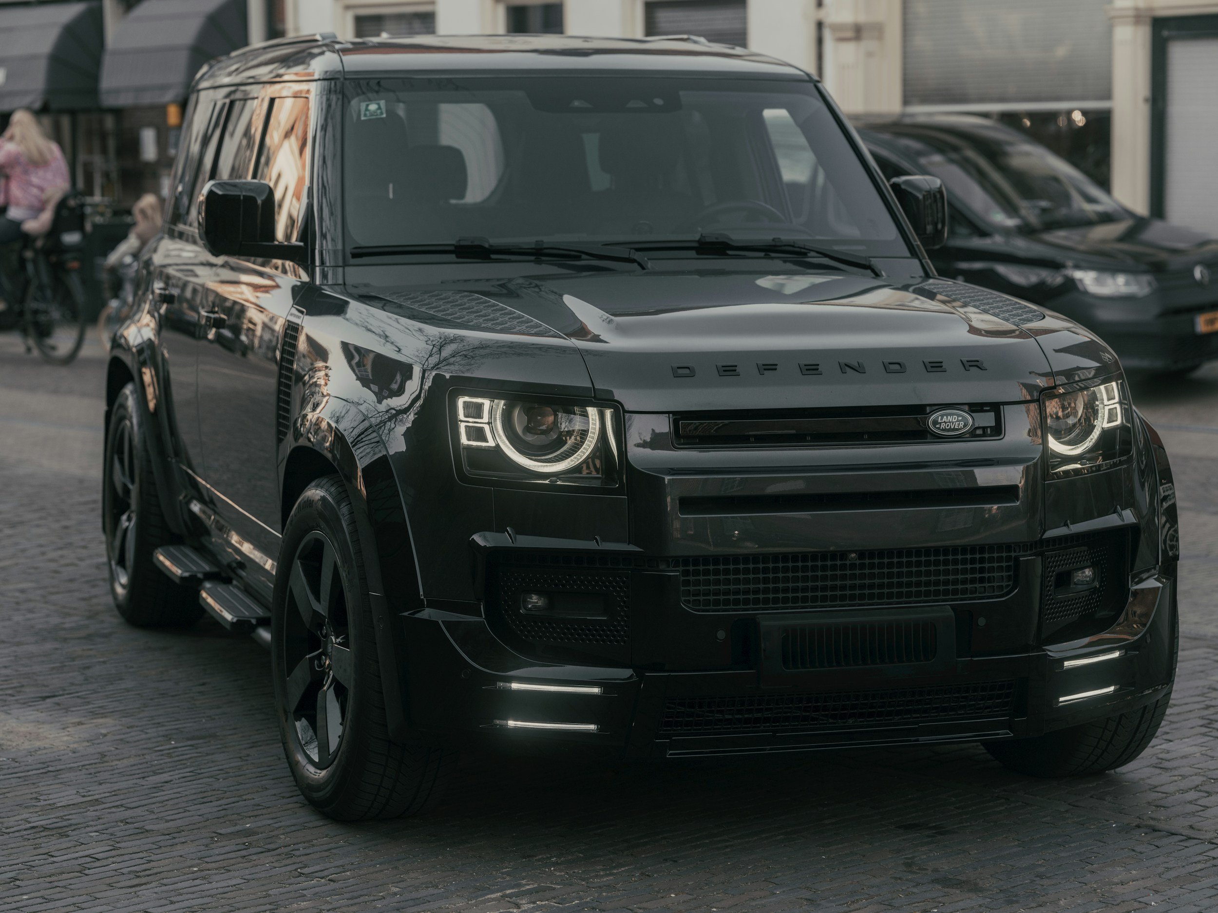 A black Land Rover Defender SUV parked on a cobblestone street in an urban area, with buildings and other cars in the background.