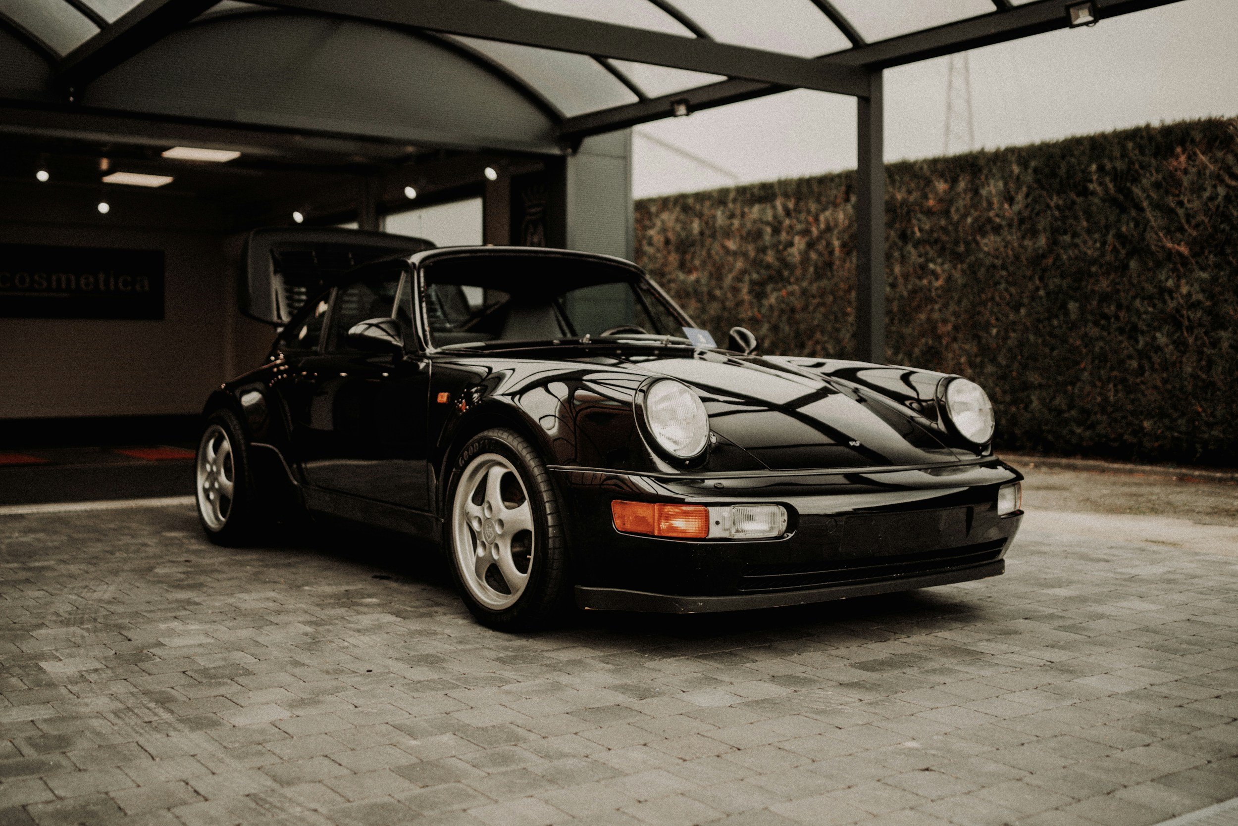 A black vintage Porsche 911 sports car parked under a shelter with a hedge in the background.