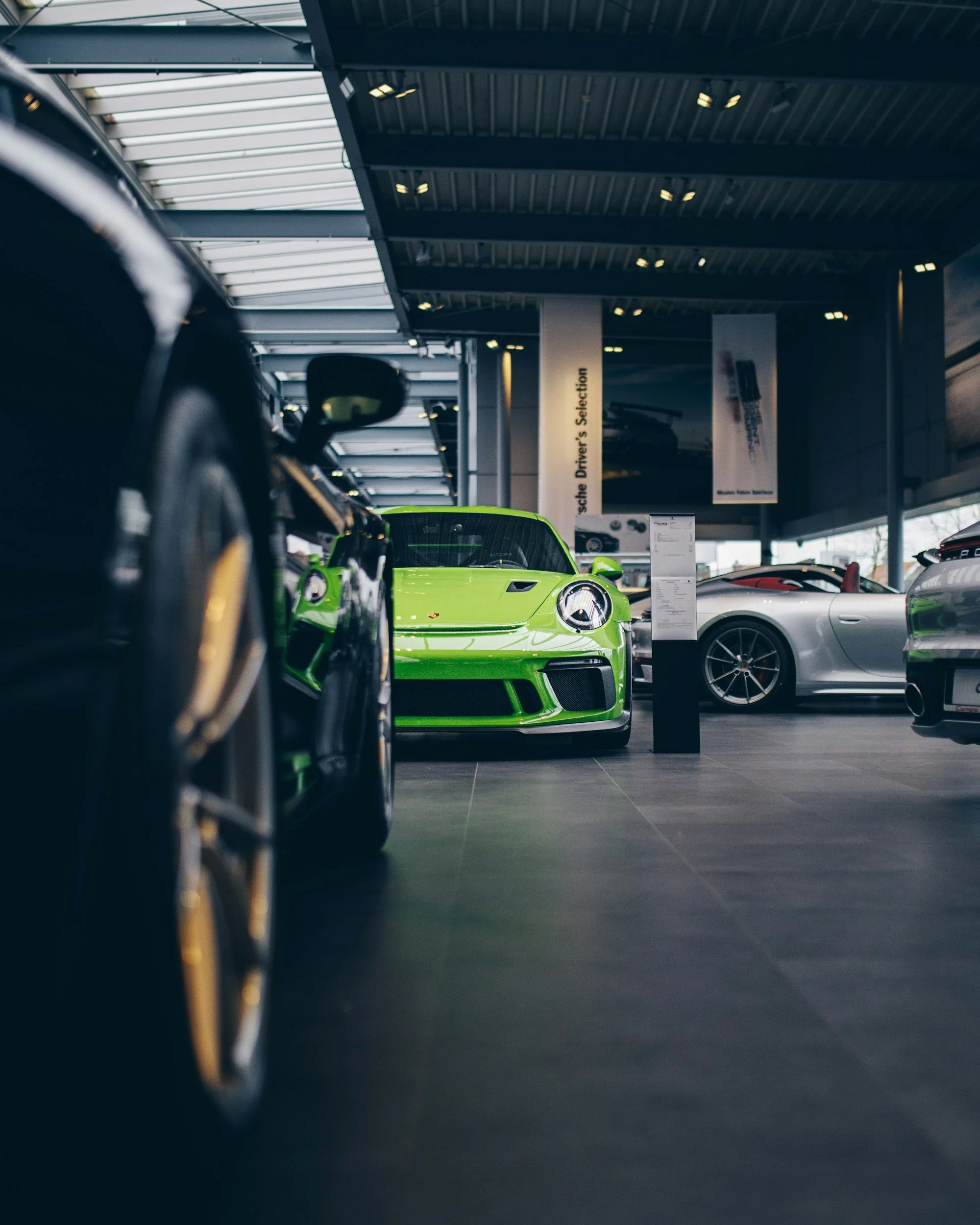 View of a green sports car inside a car dealership with other vehicles, ceiling lights, and banners in the background.