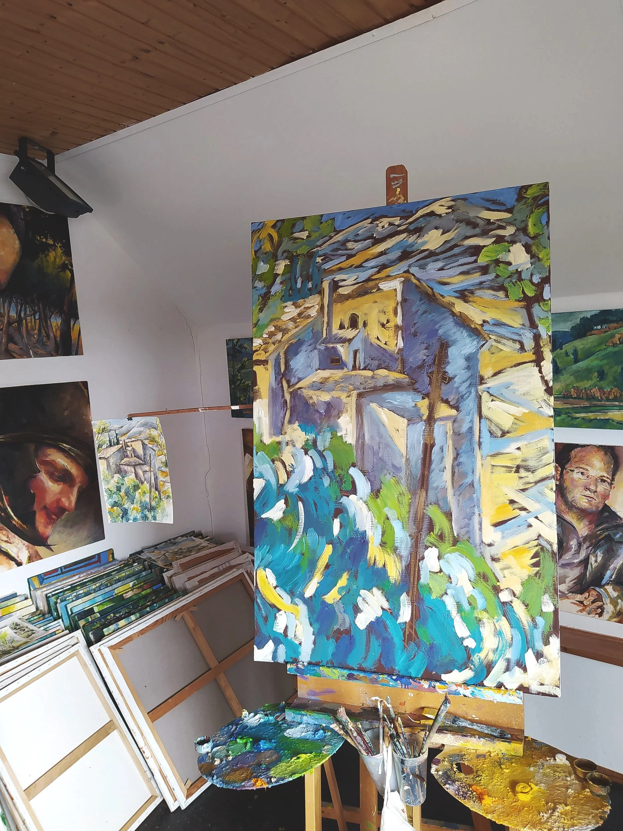 Ein Kunstraum mit mehreren Gemälden, Leinwänden und Malutensilien. Im Vordergrund ein großes, farbenfrohes Gemälde, daneben eine Palette mit bunten Farben und Pinsel. An der Wand hängen verschiedene Kunstwerke.