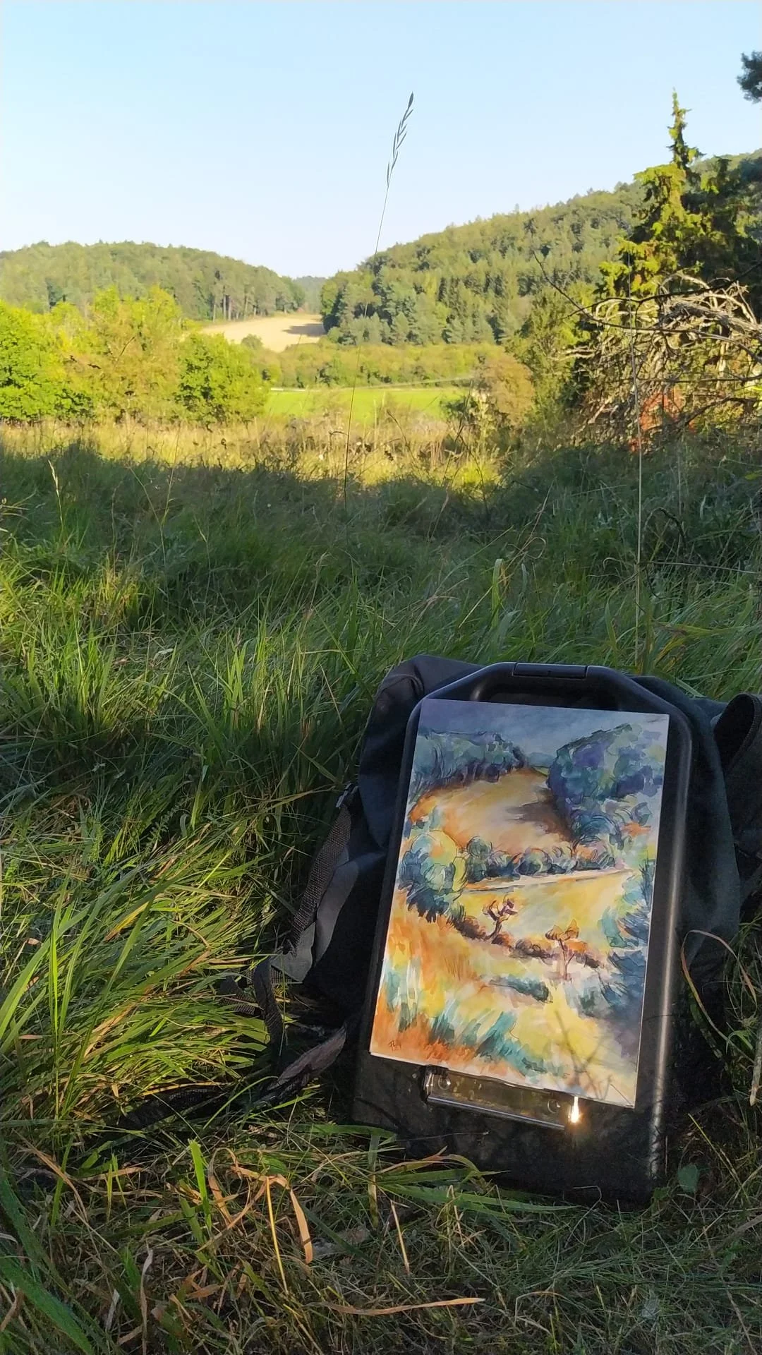 Landschaft mit Bäumen und Hügeln bei Sonnenlicht, im Vordergrund eine Tasche mit einer Malerei auf Papier, die einen Fluss und Bäume zeigt.