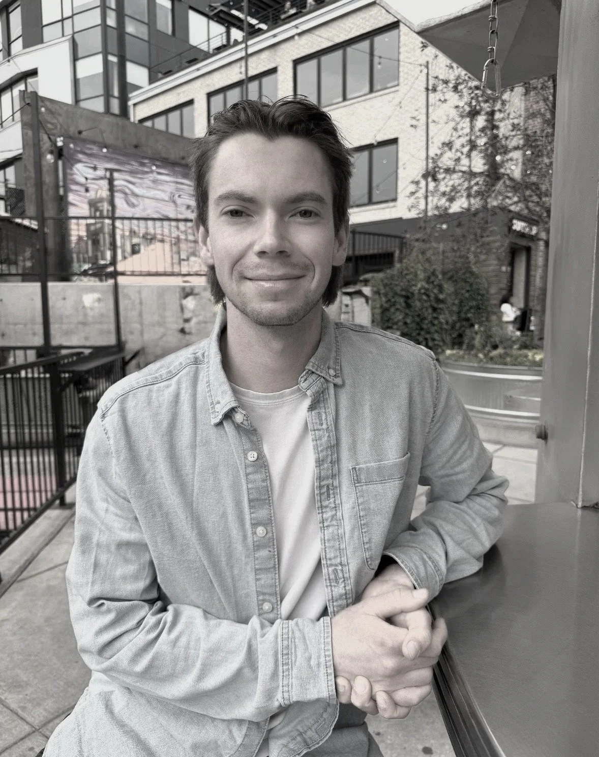 A young man with dark hair and light skin, smiling while sitting outdoors in an urban setting with buildings, trees, and a patio area in the background.
