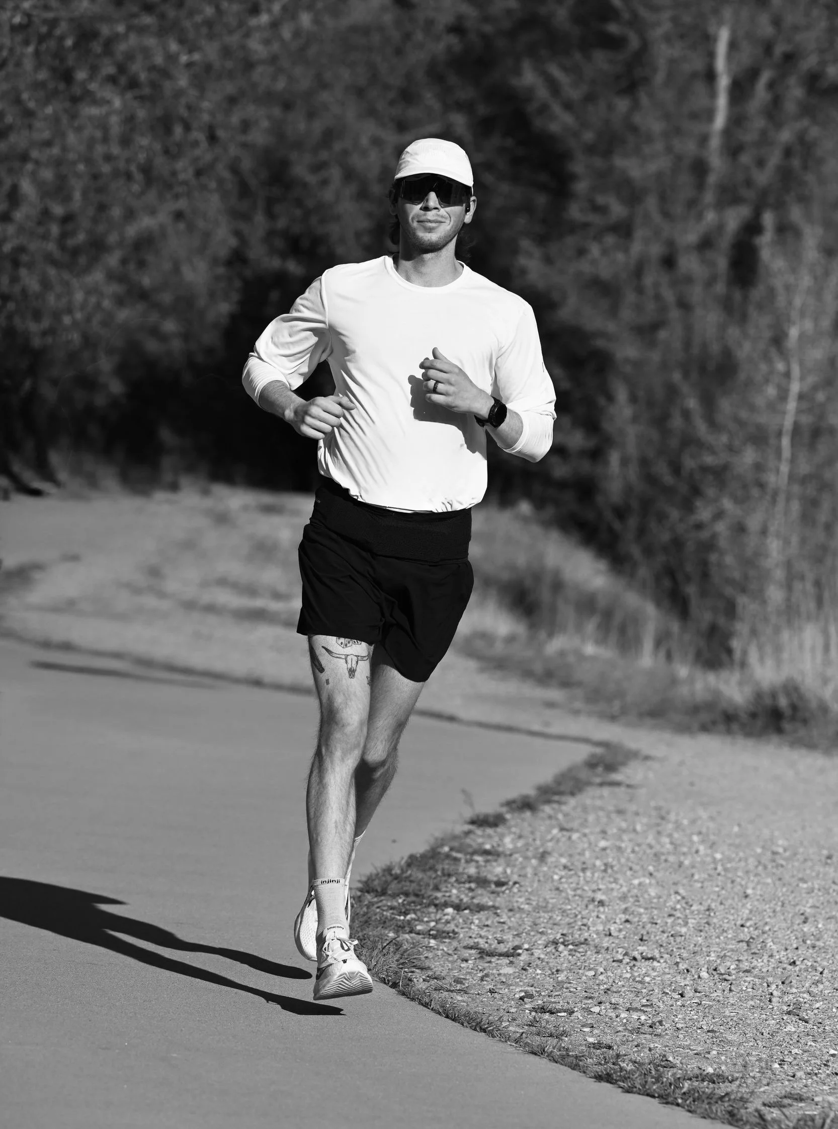 A man running outdoors in a park or trail, wearing sunglasses, a white cap, a long-sleeve shirt, black shorts, white running shoes, and a watch, with trees and a paved path in the background.
