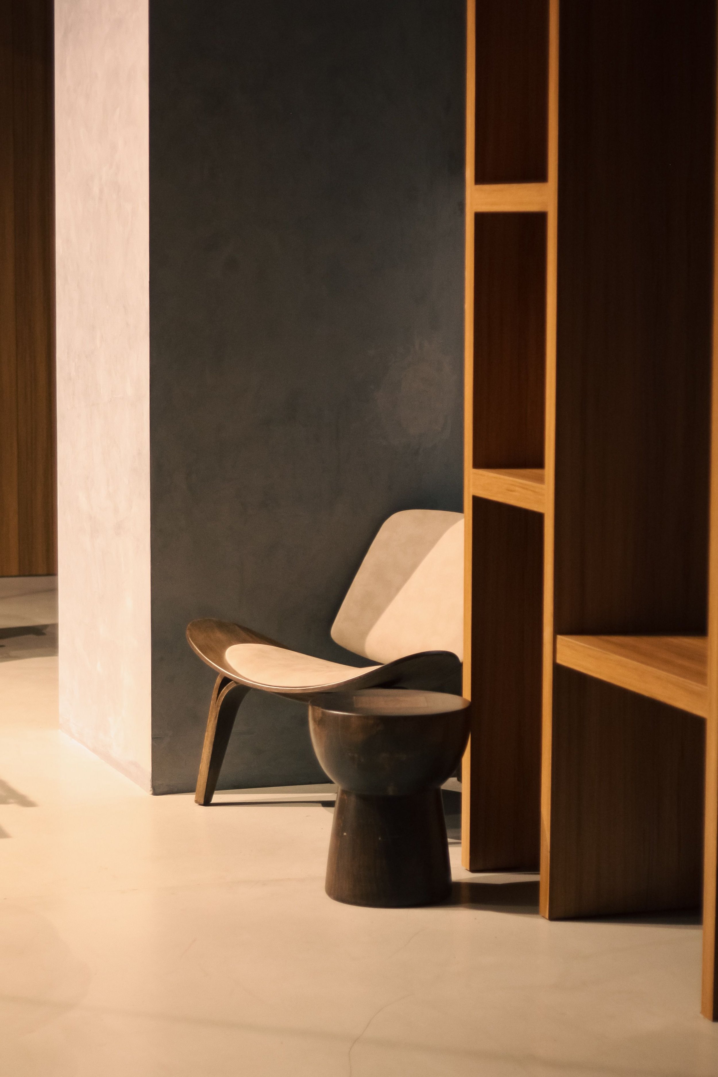 Interior space with modern furniture, including a wooden chair, a dark wooden stool, and a built-in wooden shelving unit, with a dark-colored wall background.