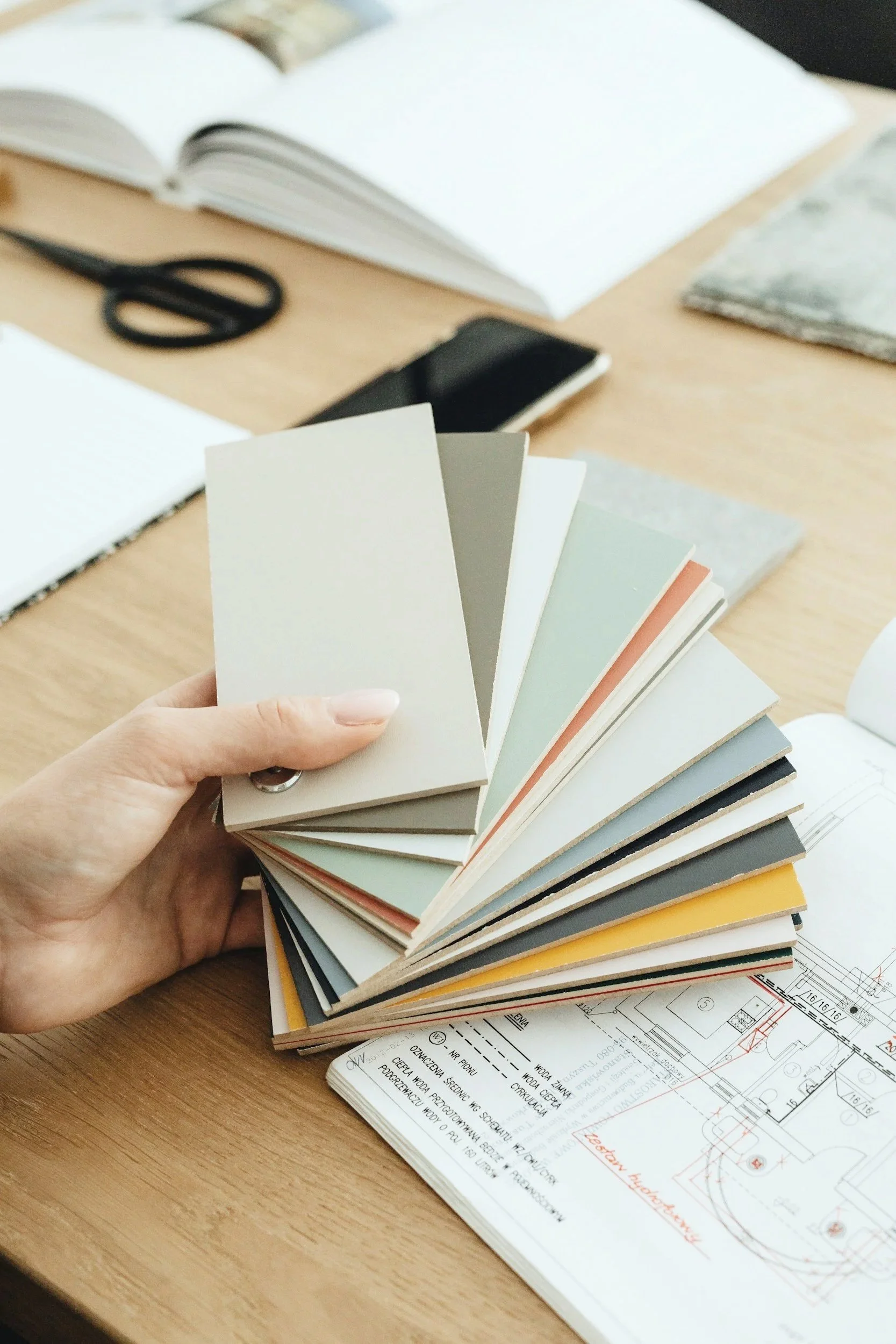 A person's hand holds a color swatch fan deck in front of a wooden table. The table also has architectural blueprints, an open notebook, and a pair of scissors.
