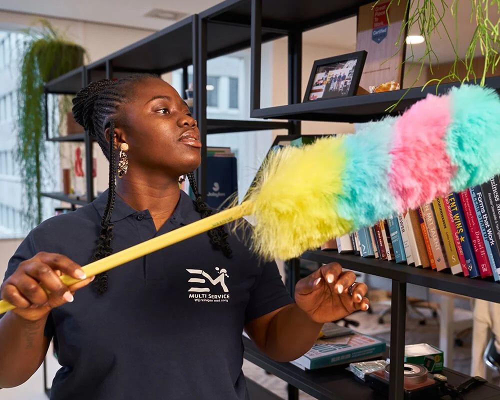 Een vrouw in een donkerblauw shirt met logo veegt stof van een boekenplank met een kleurrijke plumeau, in een kantoorruimte.