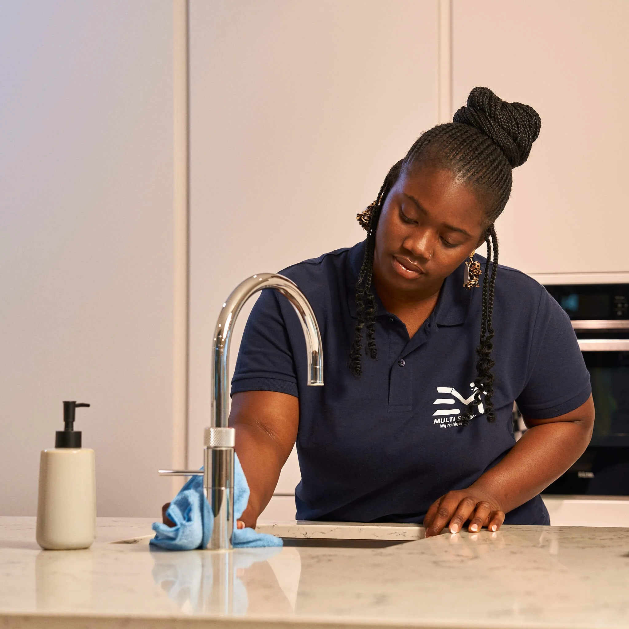 Een vrouw met gevlochten haar en een blauwe polo die op een keukenbalie werkt met schoonmaakproducten, een witte zeepdispenser en een natte doek.
