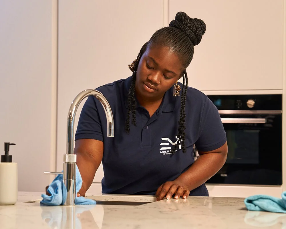 Vrouw wast een keukenaanrecht. Ze draagt een navyblauwe polo met logo en heeft haar haar in een hoge bol opgestoken.