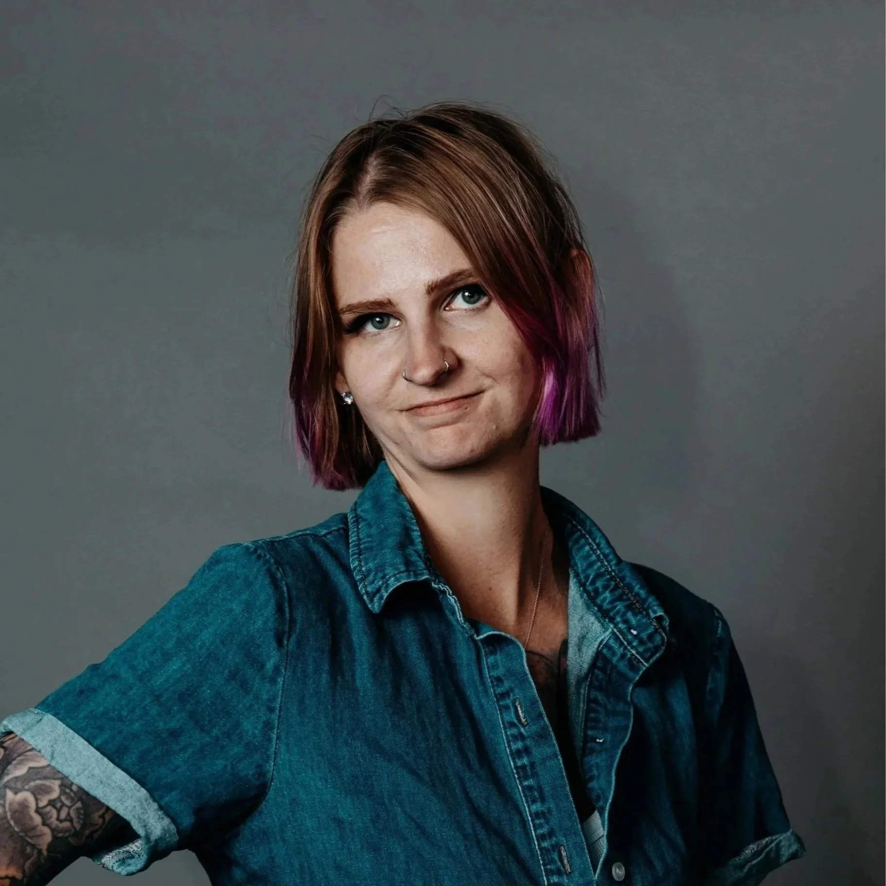 Portrait of a young woman with shoulder-length brown hair with purple tips, wearing a denim jacket and earrings, smiling slightly against a plain gray background.