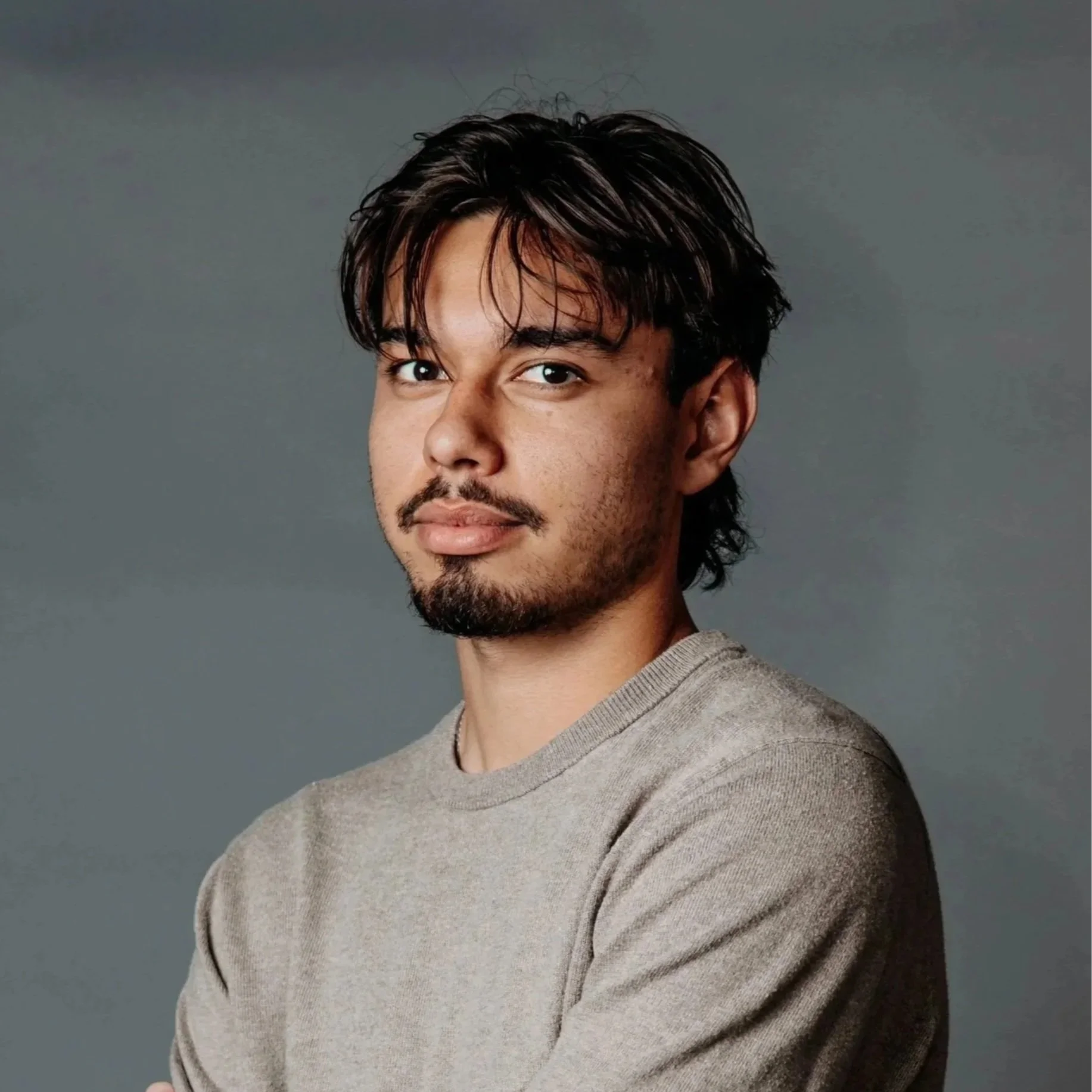 A young man with dark hair and facial hair wearing a gray sweater, standing against a dark gray background.