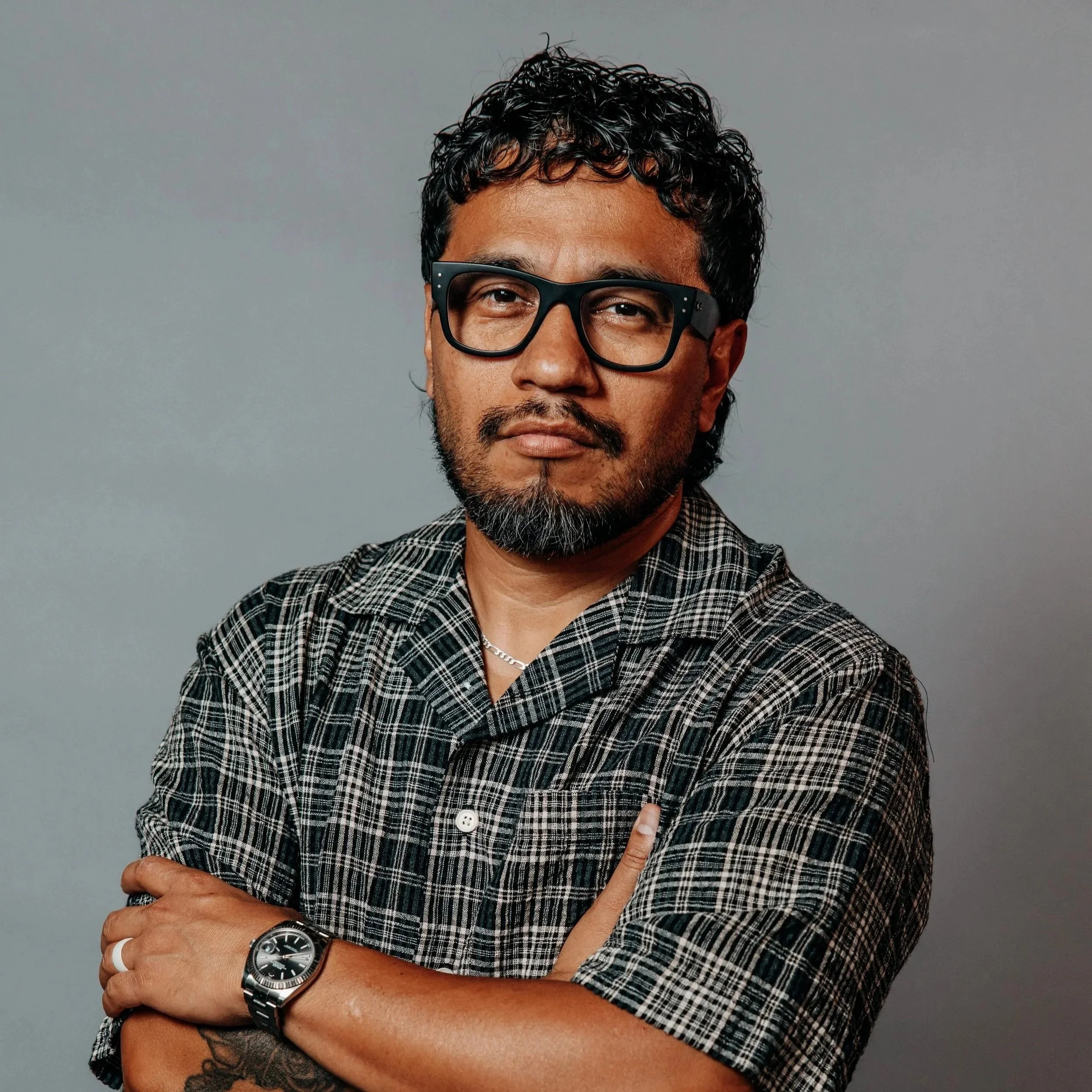 Close-up of a man wearing black glasses, a checkered shirt, a wristwatch, and pointing to his chest with one hand, against a plain gray background.