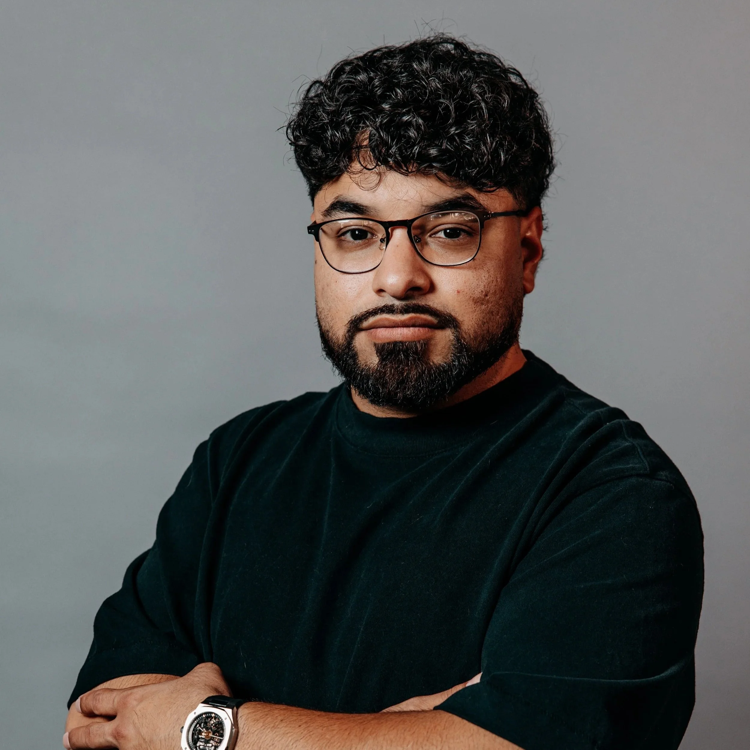 The image shows a man with curly dark hair and glasses, wearing a black T-shirt. He is crossing his arms, wearing a wristwatch, and looking directly at the camera against a plain gray background.