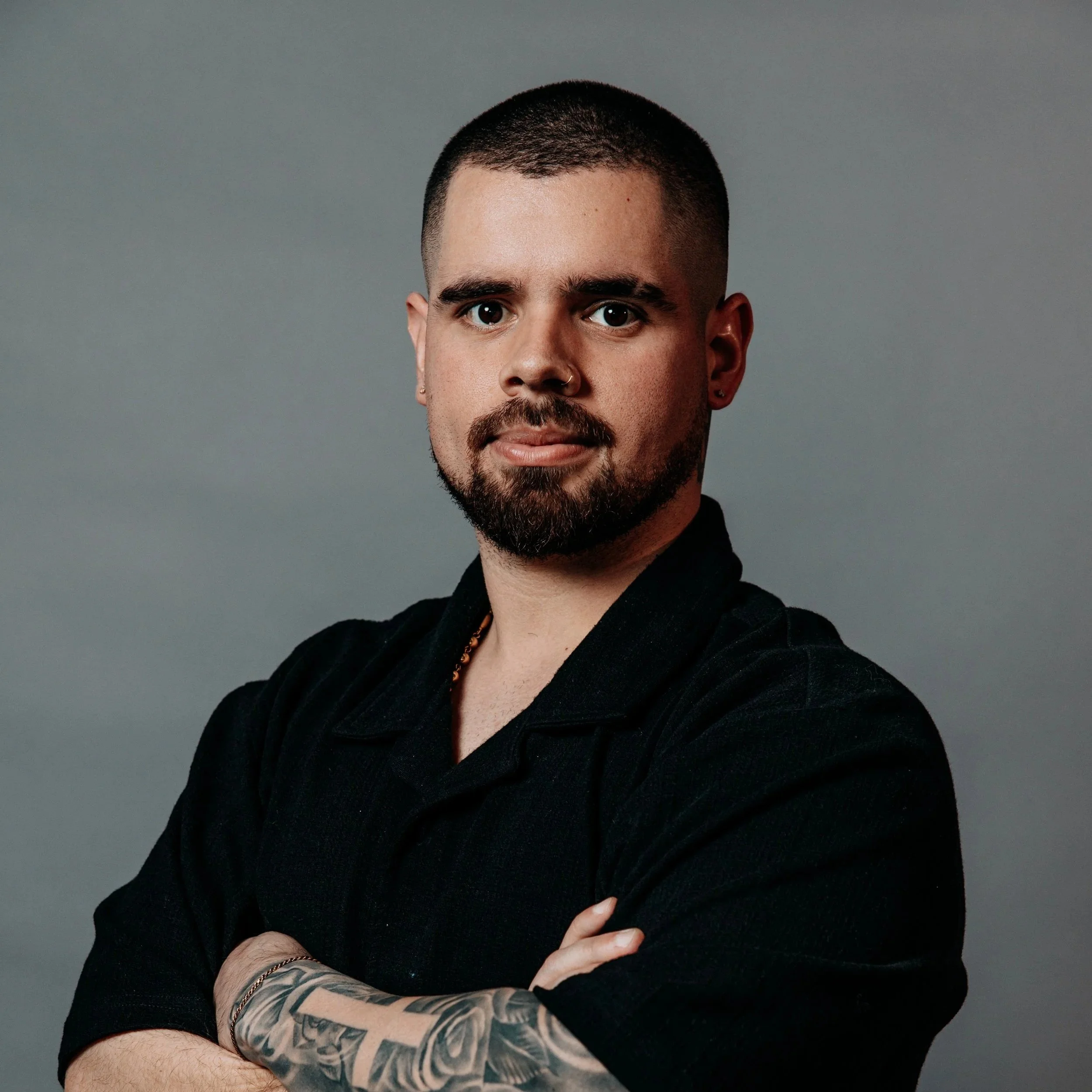 Close-up of a man with a beard and short hair, wearing a black shirt, standing against a plain gray background.
