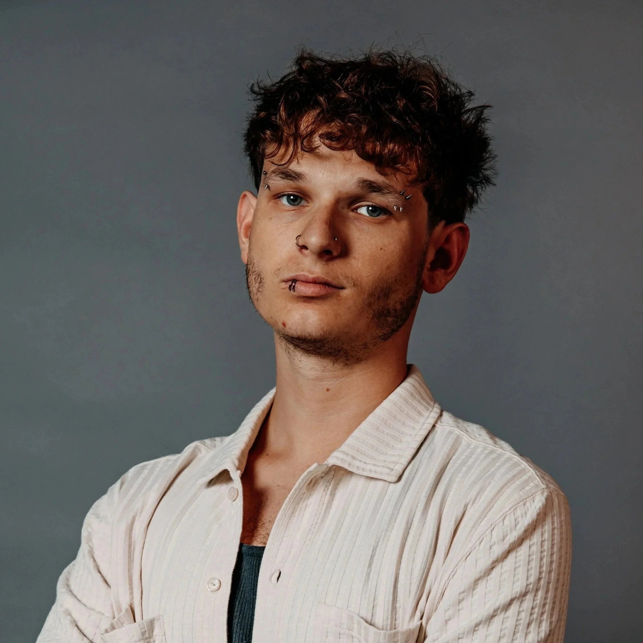 Close-up of a young man with curly brown hair, blue eyes, and multiple facial piercings, wearing a beige striped jacket over a navy shirt, against a gray background.