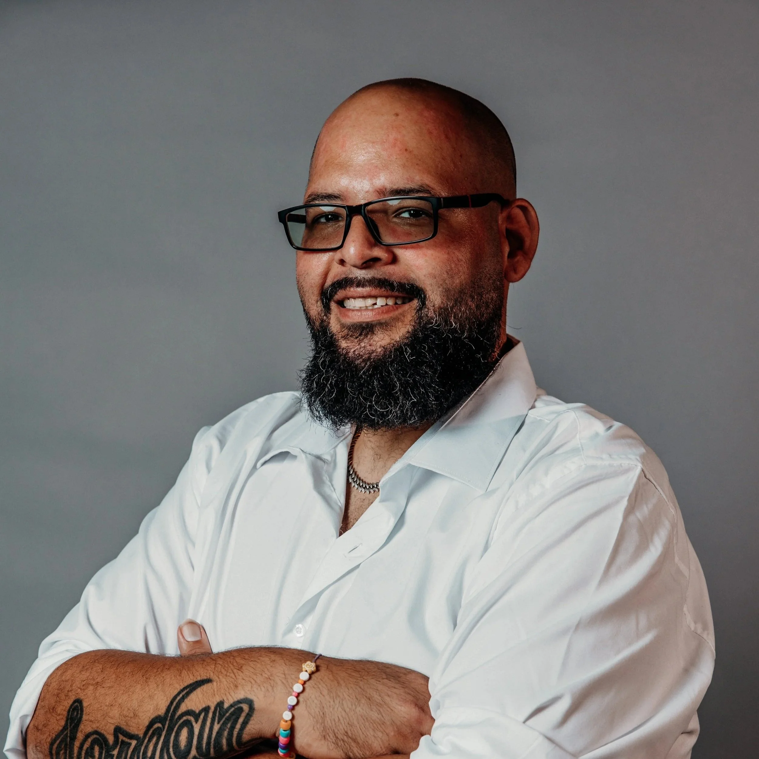 A man with glasses, a beard, and a shaved head smiling, wearing a white shirt, a colorful beaded bracelet, and a tattoo on his arm, against a plain gray background.
