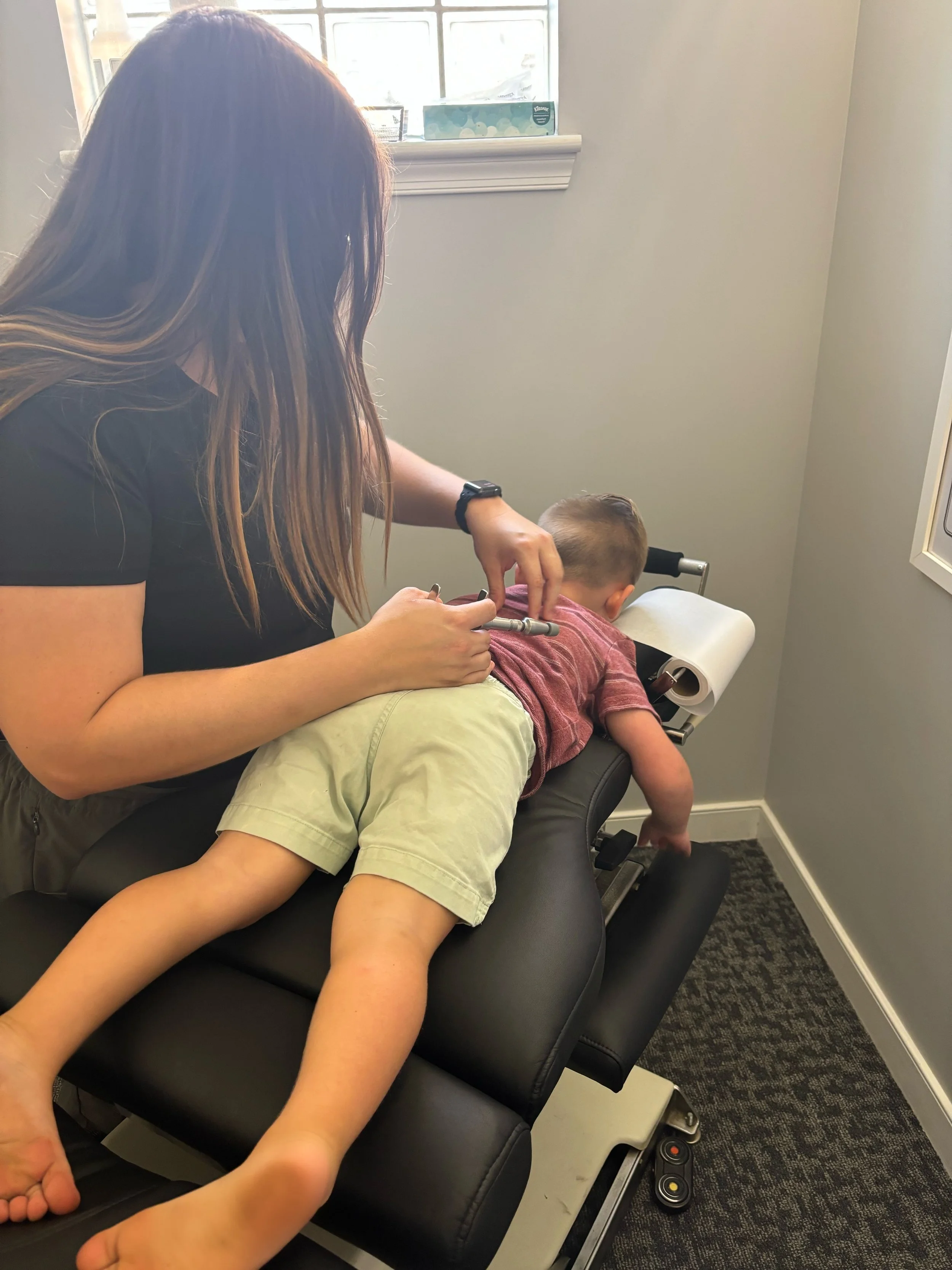A woman with long brown hair is giving a vaccination or injection to a young boy lying face down on an examination table. The boy is wearing a red striped shirt and khaki shorts, and the room has a window with partially closed blinds.