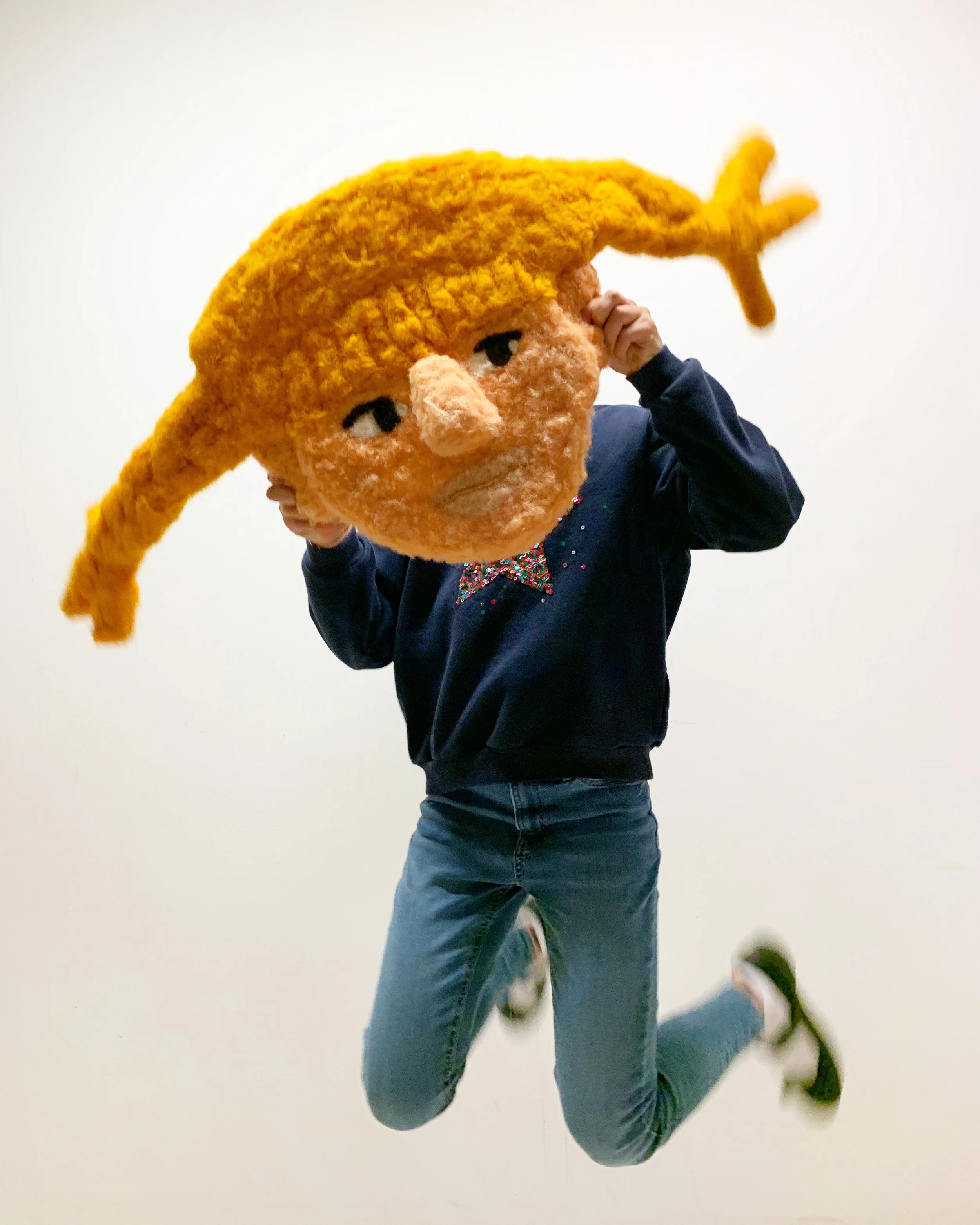 Person in a dark jacket and blue jeans holding a large wool felted toy head, in front of a white background.