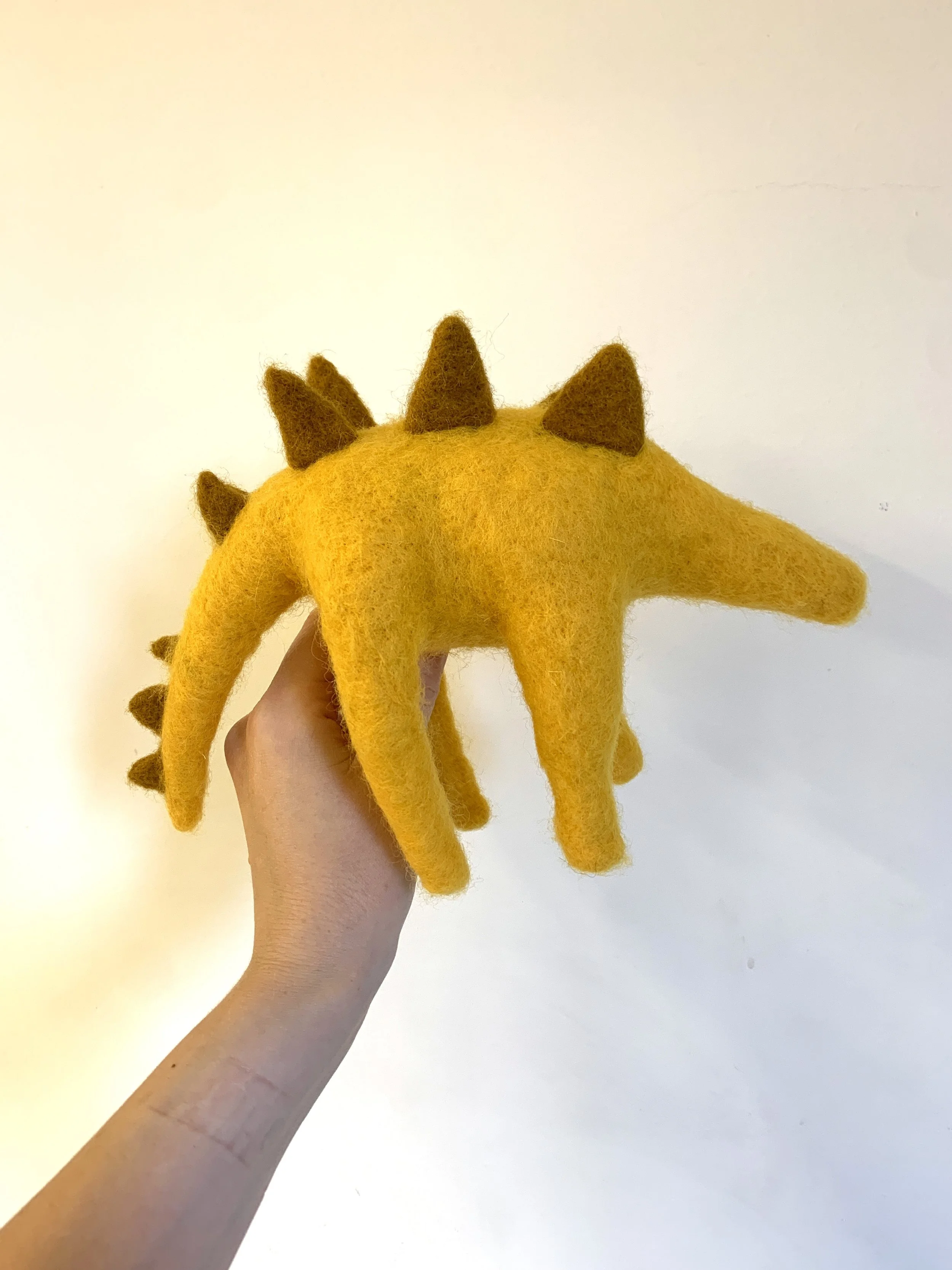 Felt plush toy of a yellow dinosaur with brown spikes along its back and tail, held by a person's hand against a plain light-colored background.