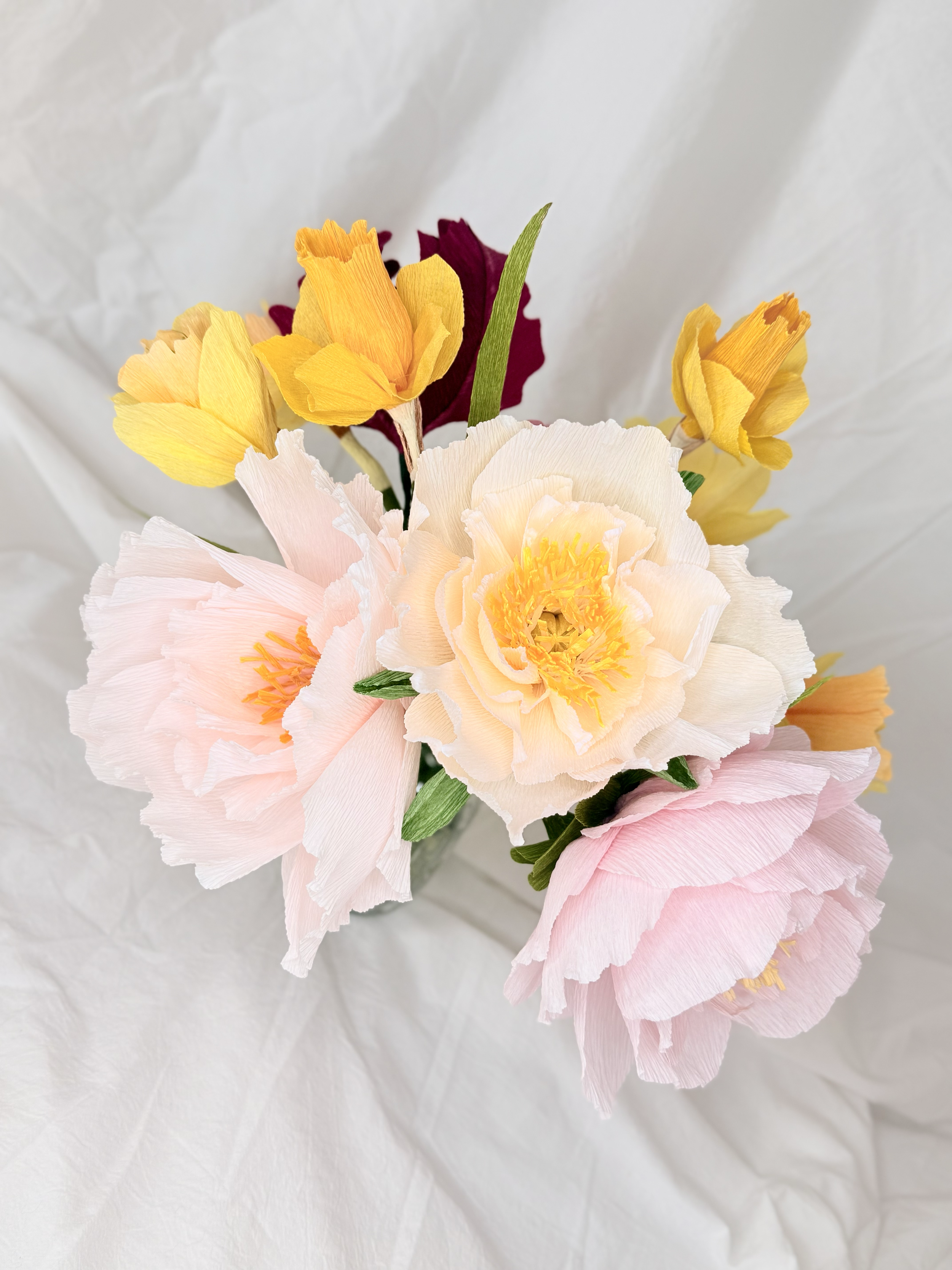 A bouquet of handmade paper flowers with cream, pink, yellow, and dark red blossoms, placed on crumpled white paper.