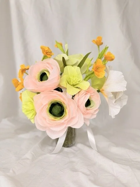 Artificial flower arrangement with pink, yellow, white, and orange flowers in a glass vase against a white fabric background.