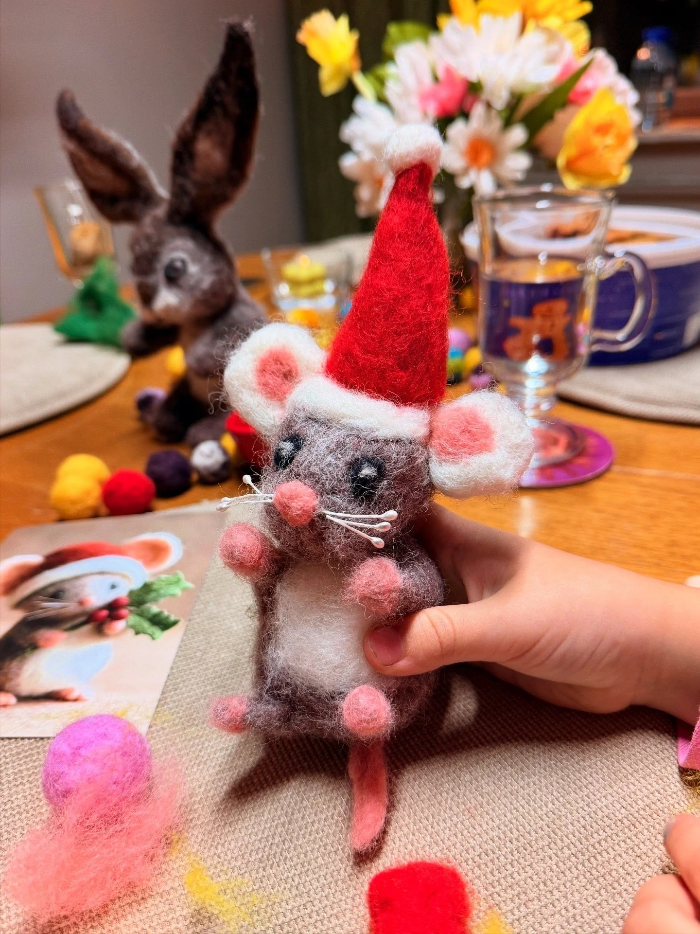 An all-felted mouse wearing a red Santa hat with white trim, held by a hand, with a decorated table in the background, including a rabbit-shaped woolen toy, colorful felt pom-poms, a vase of flowers, and a glass of water.