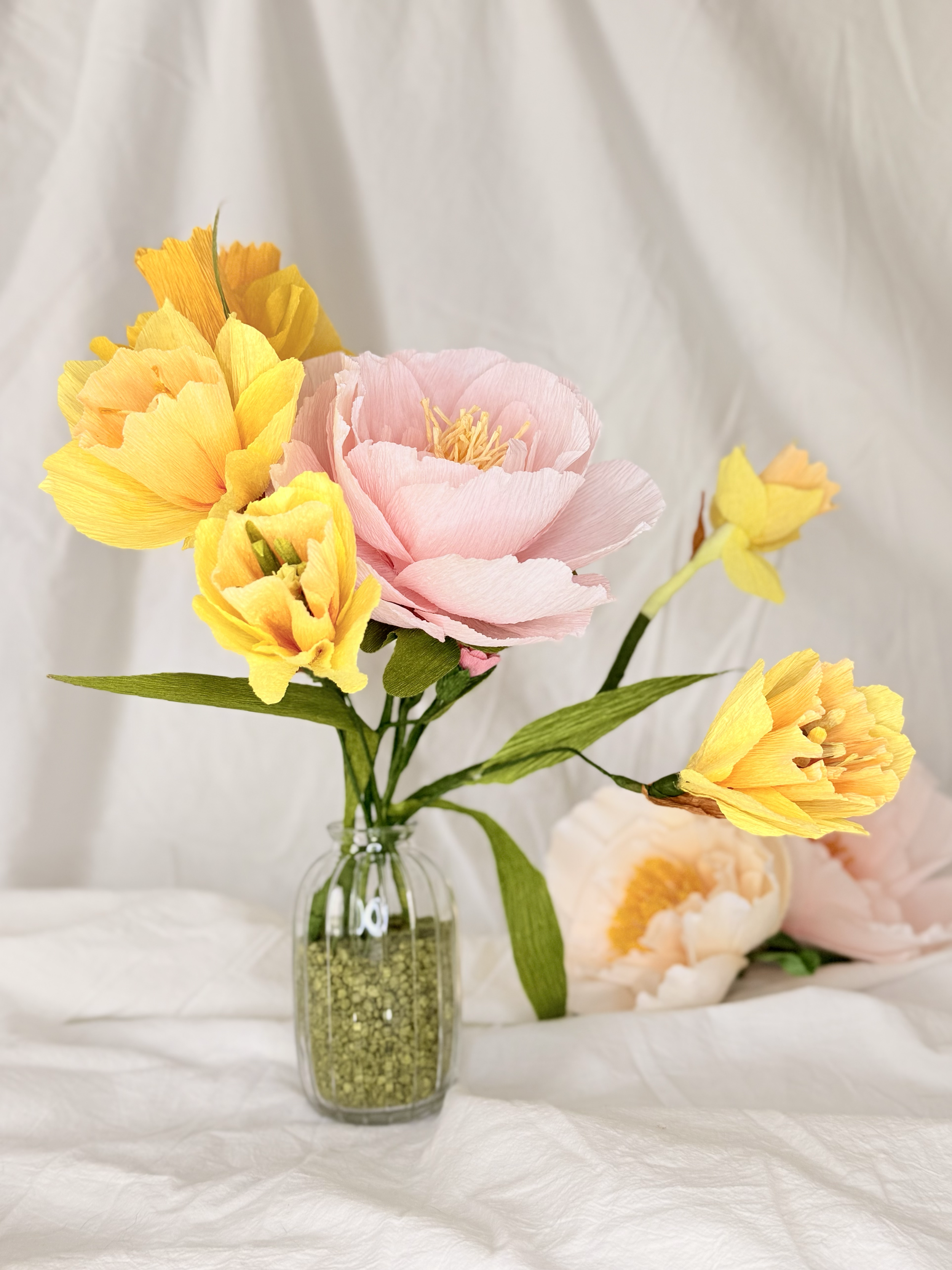 A glass vase with yellow, pink, and peach artificial flowers on a white surface with a blurred white background.