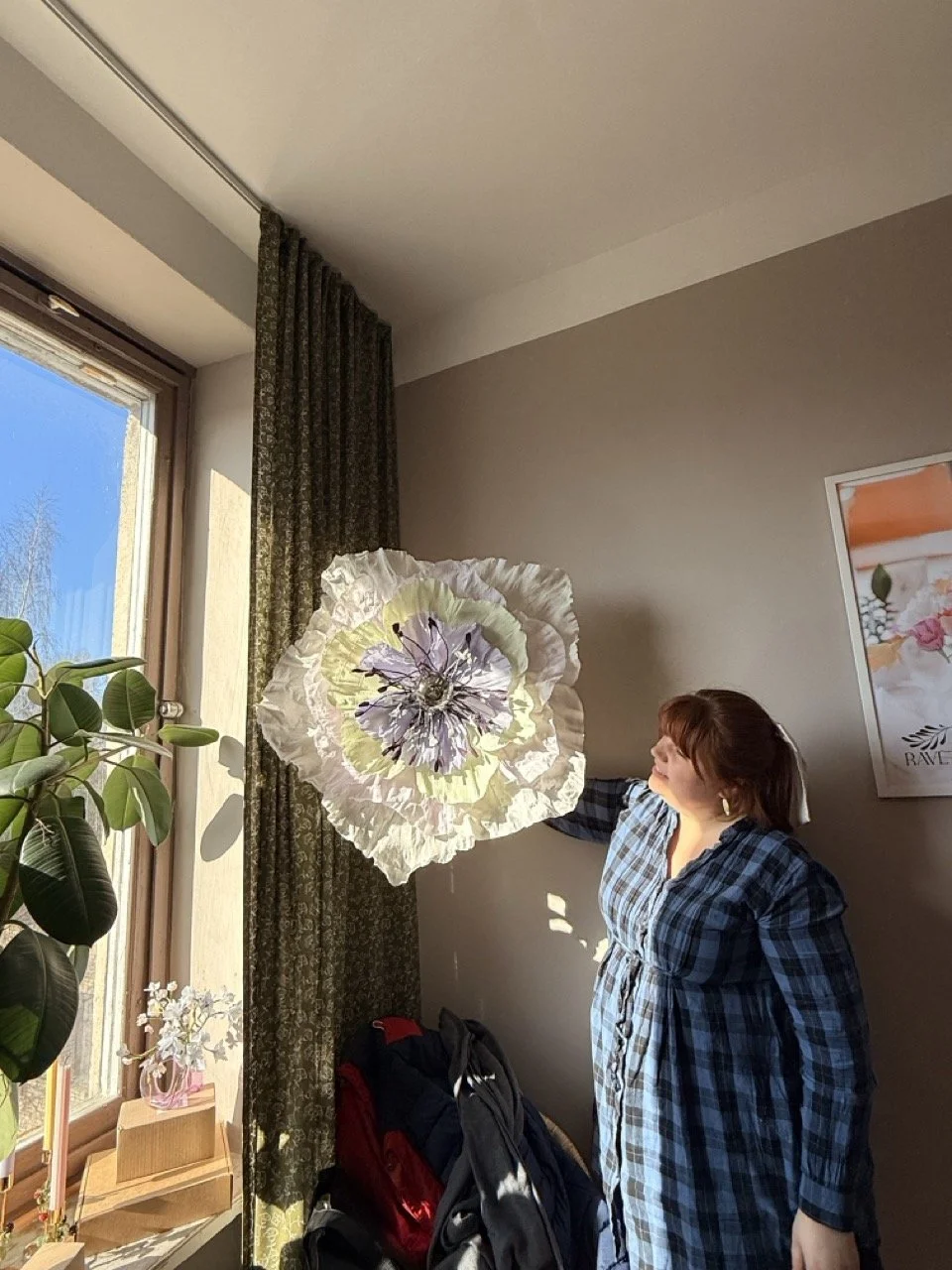 A woman in a blue plaid dress brightens a large paper flower near a window in a room with a plant and a framed picture on the wall.