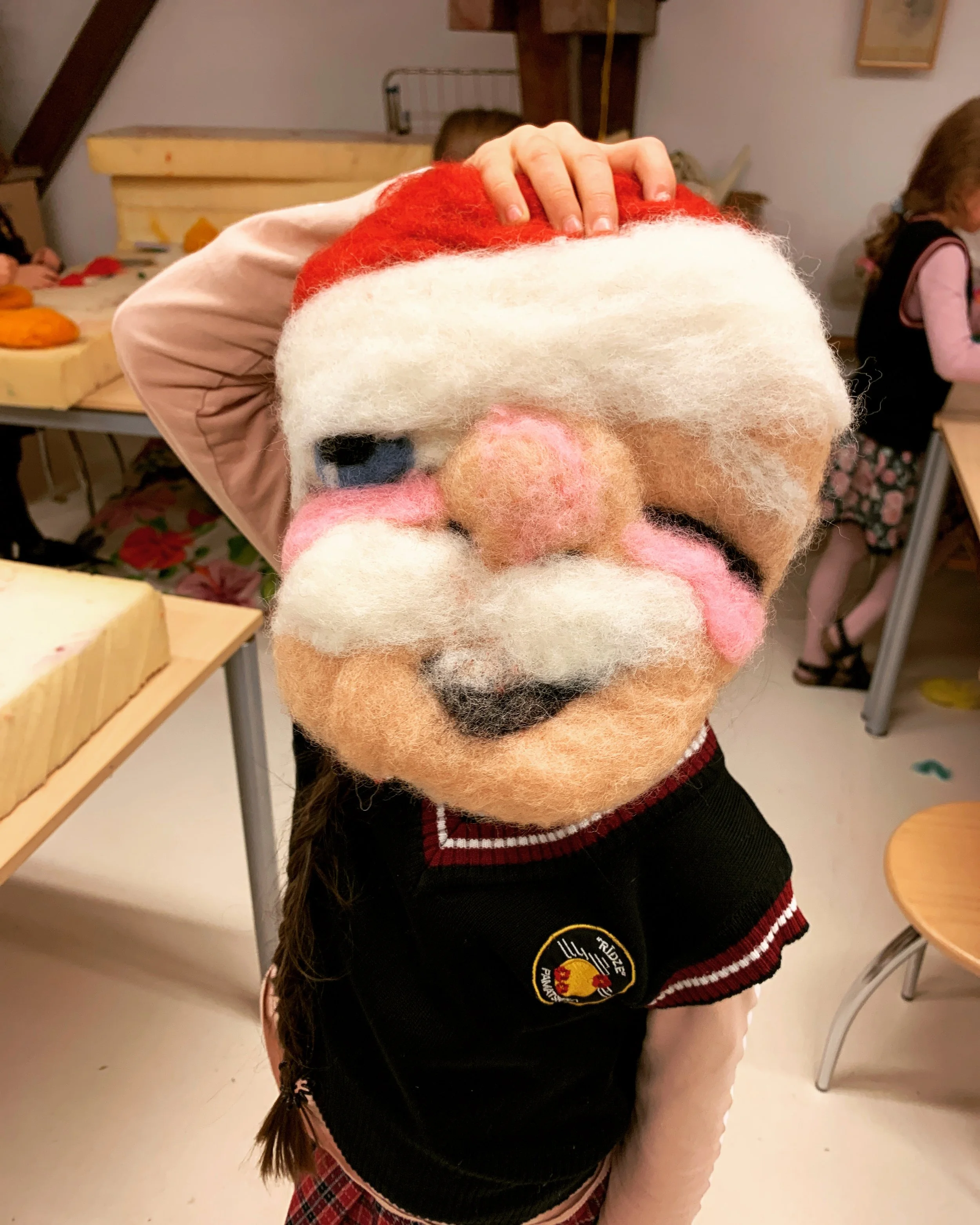 A person holding a wool felted artwork in from of their face resembling a Santa Claus, dressed in a black sweater with a patch on the front, and standing in a room with children and tables with clay objects.