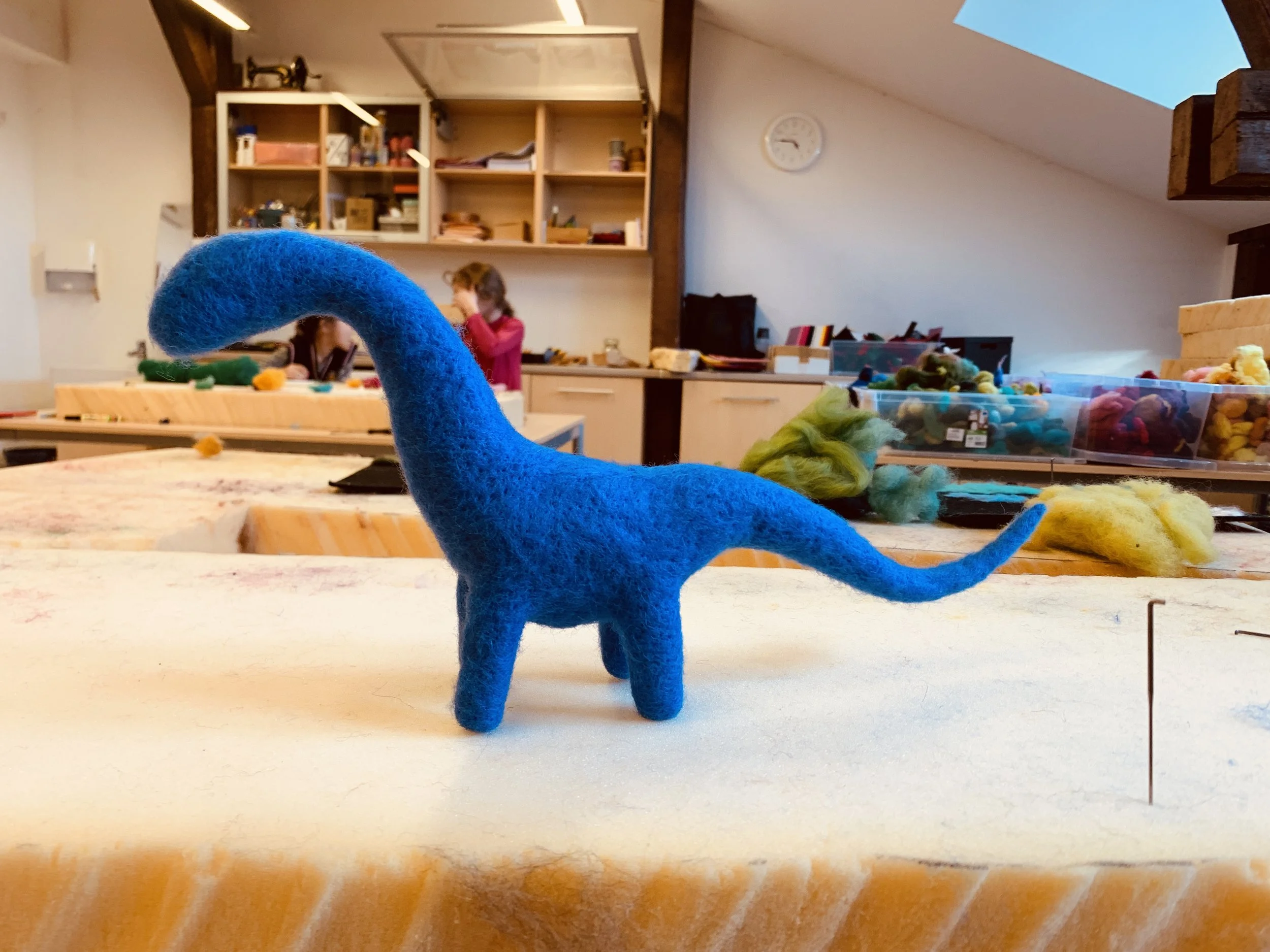 A colorful felted animal figure, possibly a dinosaur, with a long tail and a rounded body, standing on a table in a crafts room. In the background, children are working on crafts at a table with shelves filled with various art supplies and materials.