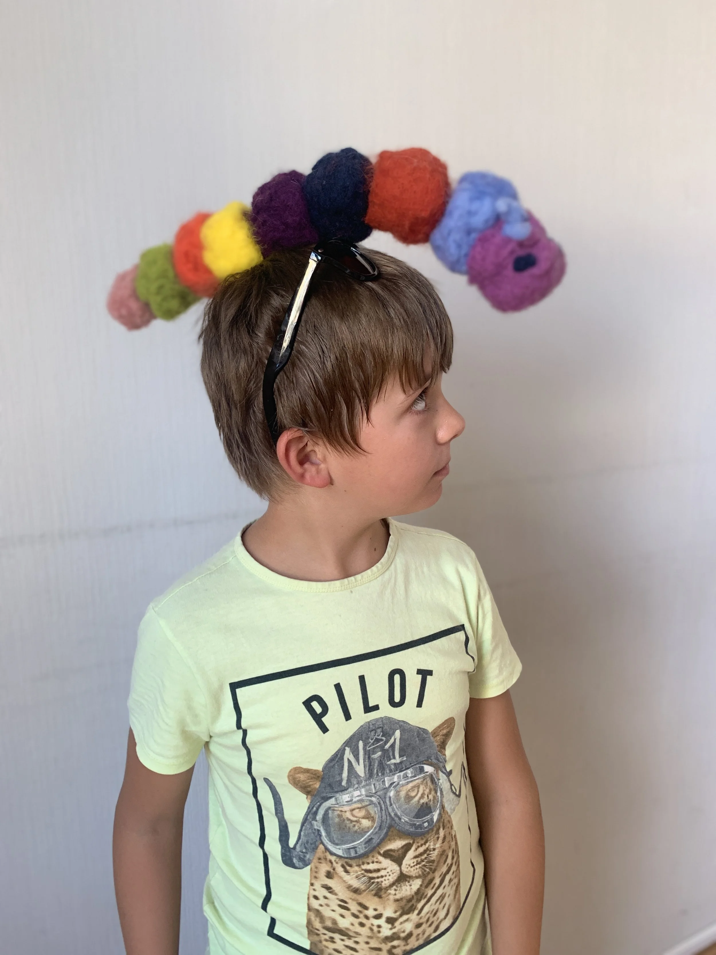 A young boy wearing glasses on his head and a yellow T-shirt with a graphic of a lion wearing aviator sunglasses and a pilot hat, standing sideways against a plain light-colored wall.