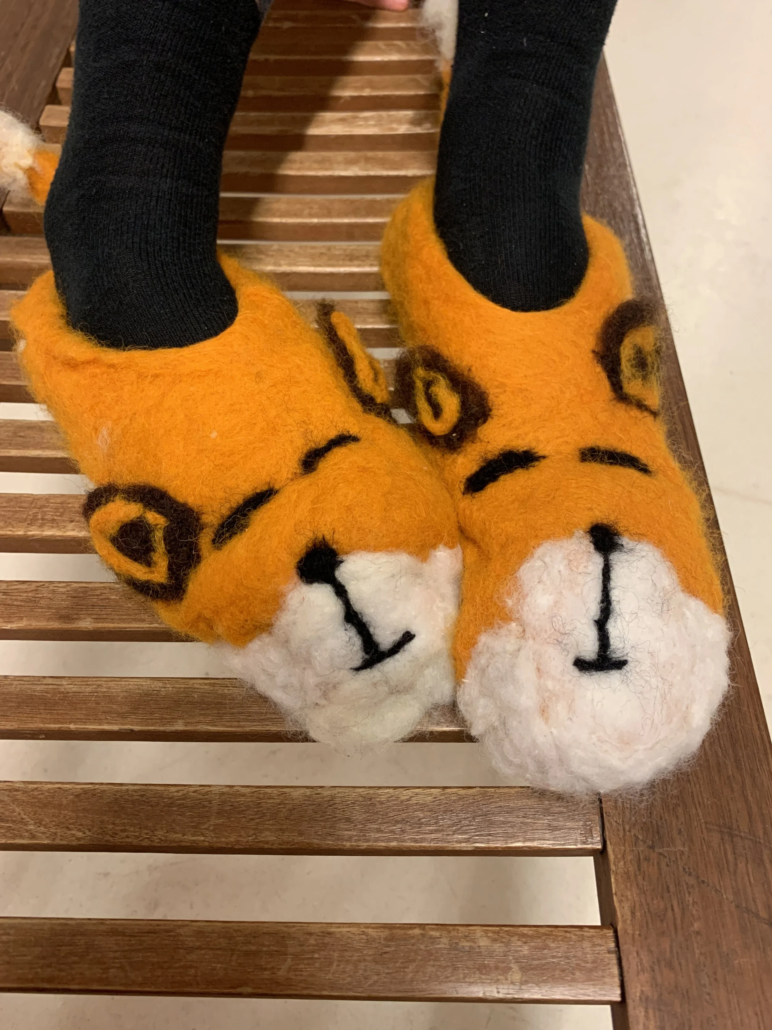 Pair of fuzzy orange slippers with tiger face design on the top, worn over black socks, placed on a wooden bench.