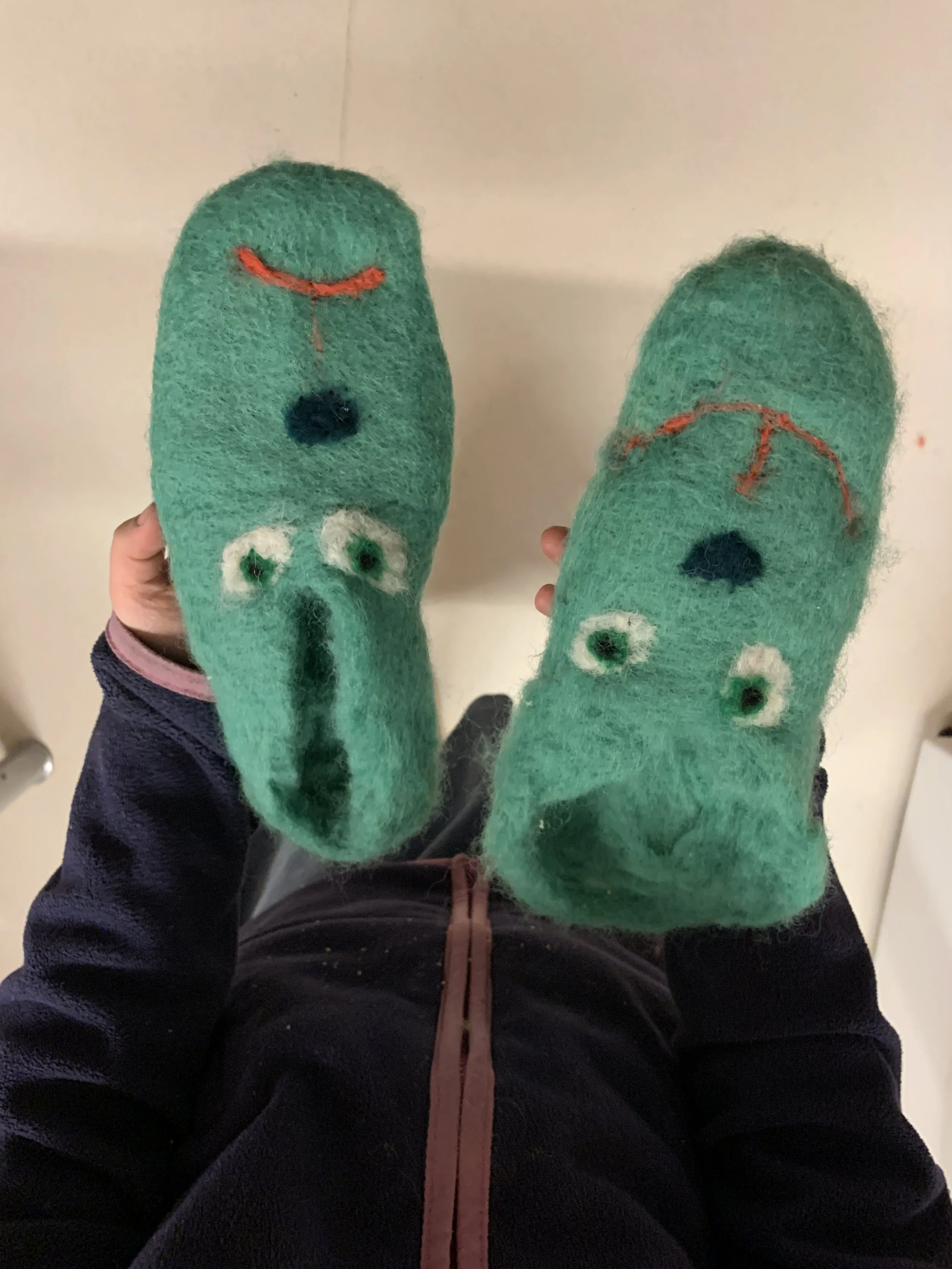 Close-up of the feet of a person wearing green fuzzy slippers with smiley faces and googly eyes, held up in front of a mirror.